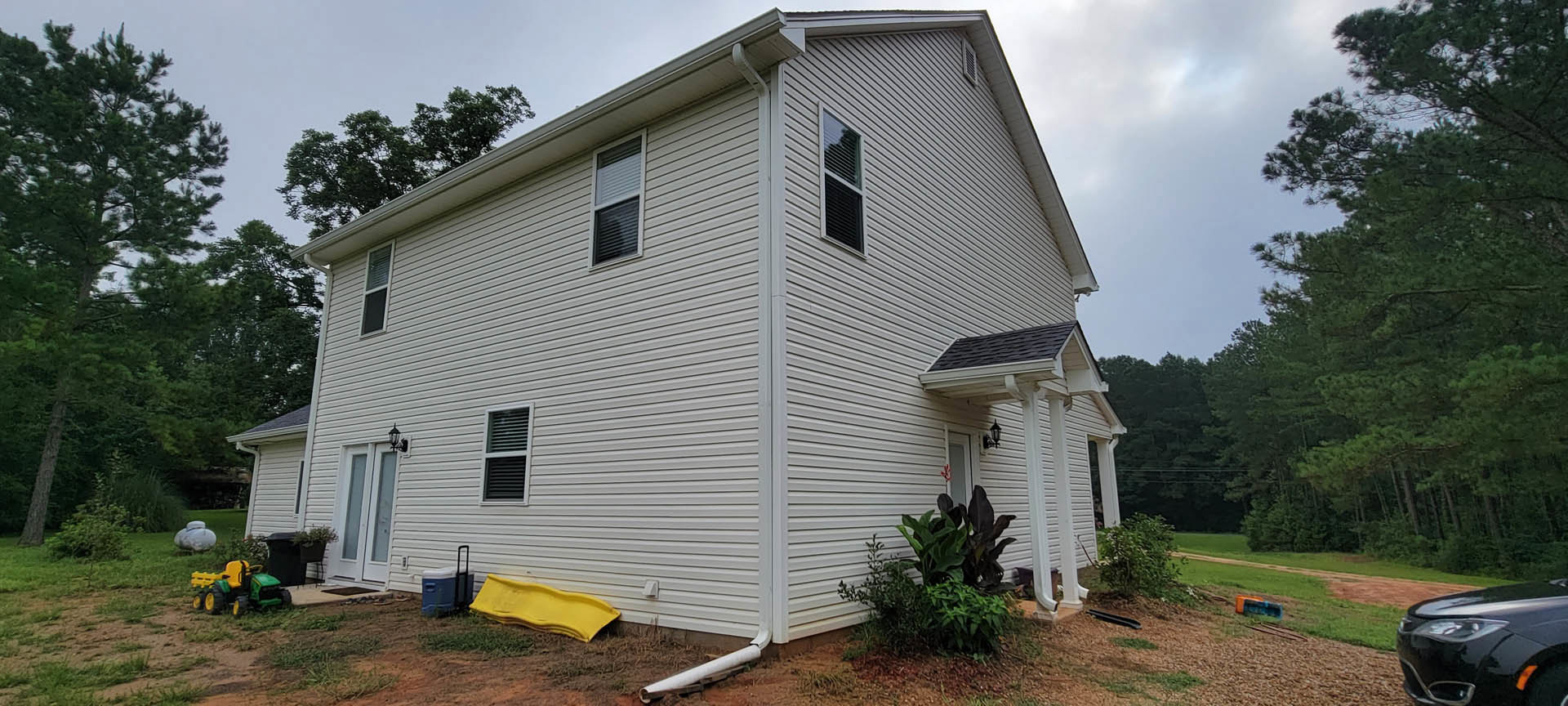 White metal roof on a modern cottage, surrounded by green grass and trees, black car parked in driveway, yellow plastic covering on ground near entry, window with closed blinds