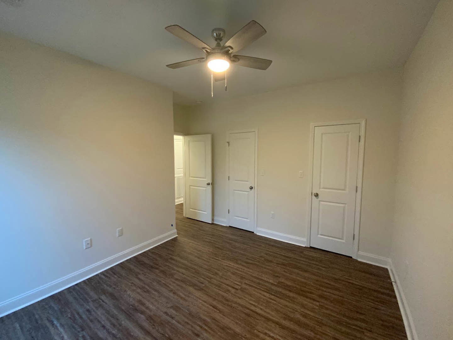 Room with white walls, wood laminate flooring, ceiling fan with light, and white door featuring a silver handle