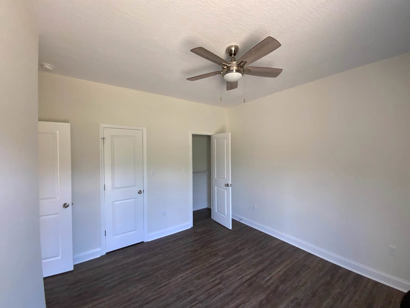 Room with white walls, wood flooring, white doors with silver handles, and a ceiling fan with light fixture
