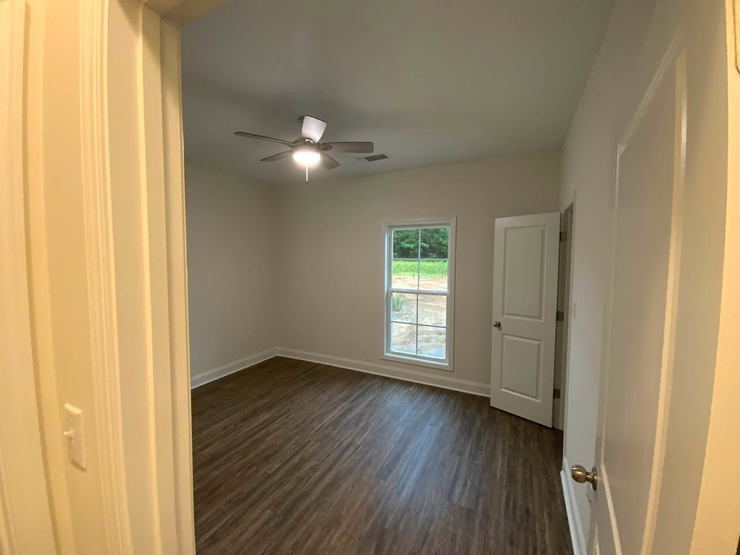 Bedroom with laminate wood flooring, white walls, ceiling fan with light, large window overlooking grassy field, white door with silver knob, small white thermostat on wall