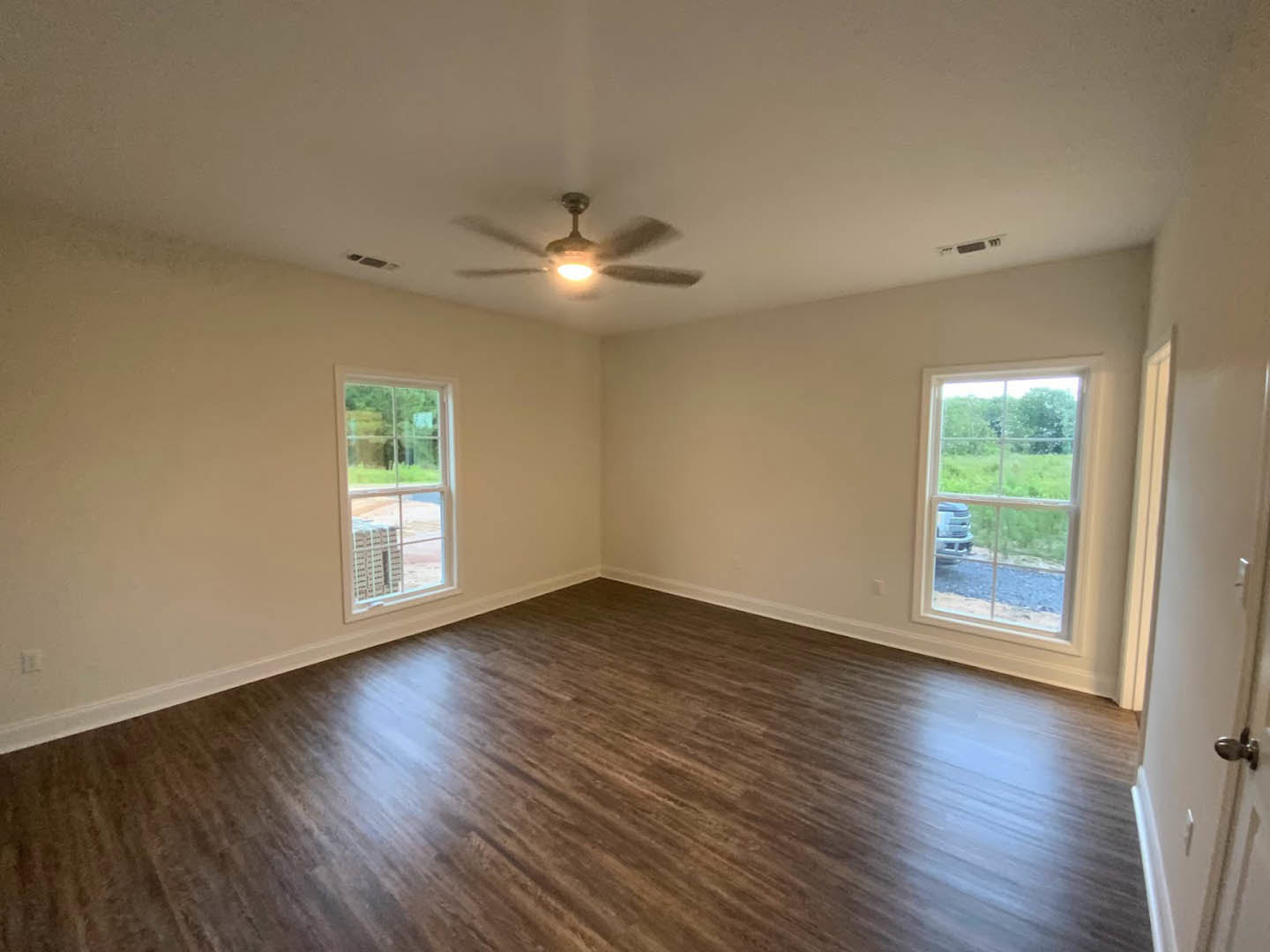 Living room with wood flooring, white plaster walls, ceiling fan with light, and large windows showing a parked car and grassy yard with trees