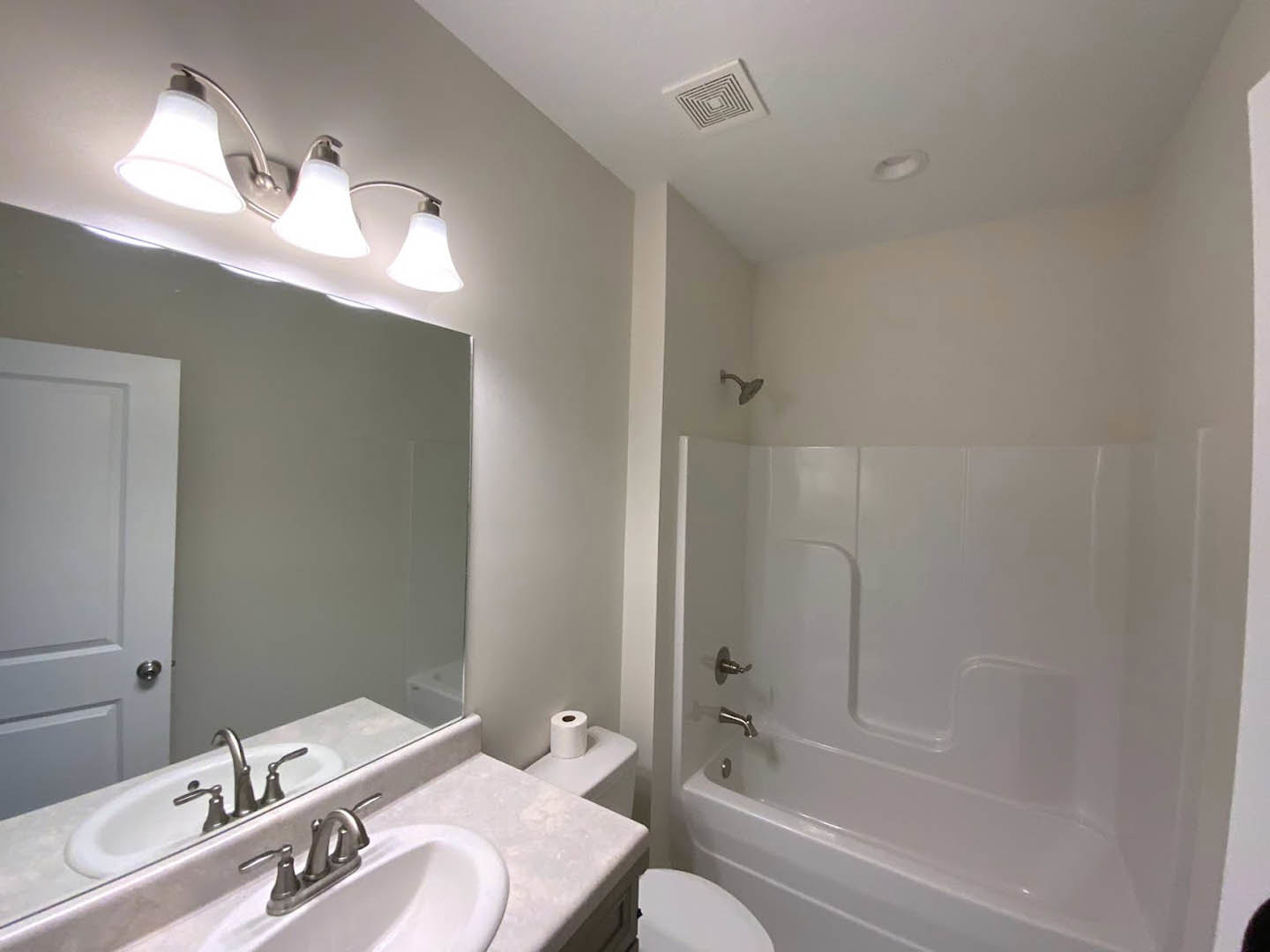 Bathroom with white tile walls, built-in bathtub, rectangular mirror above a modern sink and chrome faucet, three-light fixture, ceiling vent, and toilet paper holder
