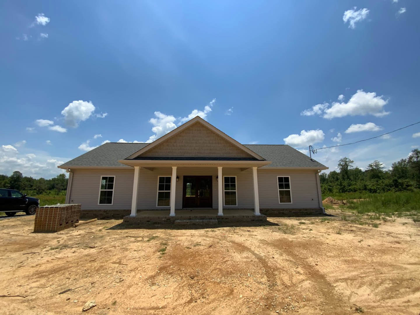 Two-story house with gray roof and white columns, double front doors with glass windows, dirt patch in front yard, black car parked on driveway, blue sky with scattered clouds