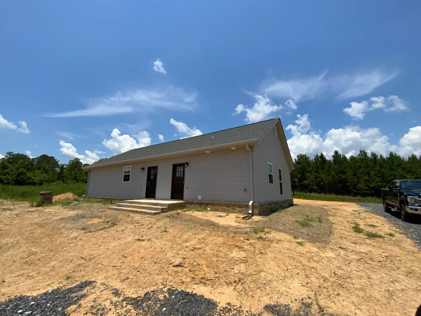 Partially built house with exposed framing, front porch, gravel driveway, black pickup truck, dirt yard, and surrounding trees under a cloudy sky