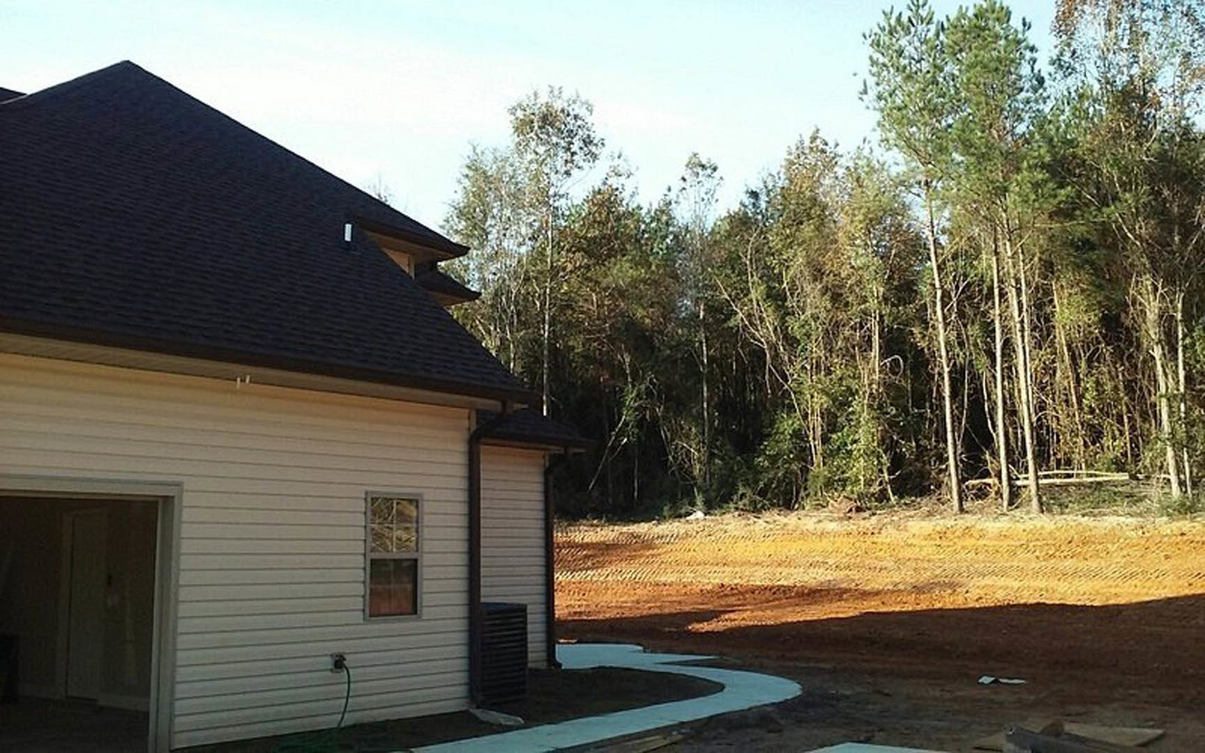 Two-story cottage with light siding, black shuttered windows, attached garage, and a dirt path leading through the backyard; mature trees and blue sky in the background.