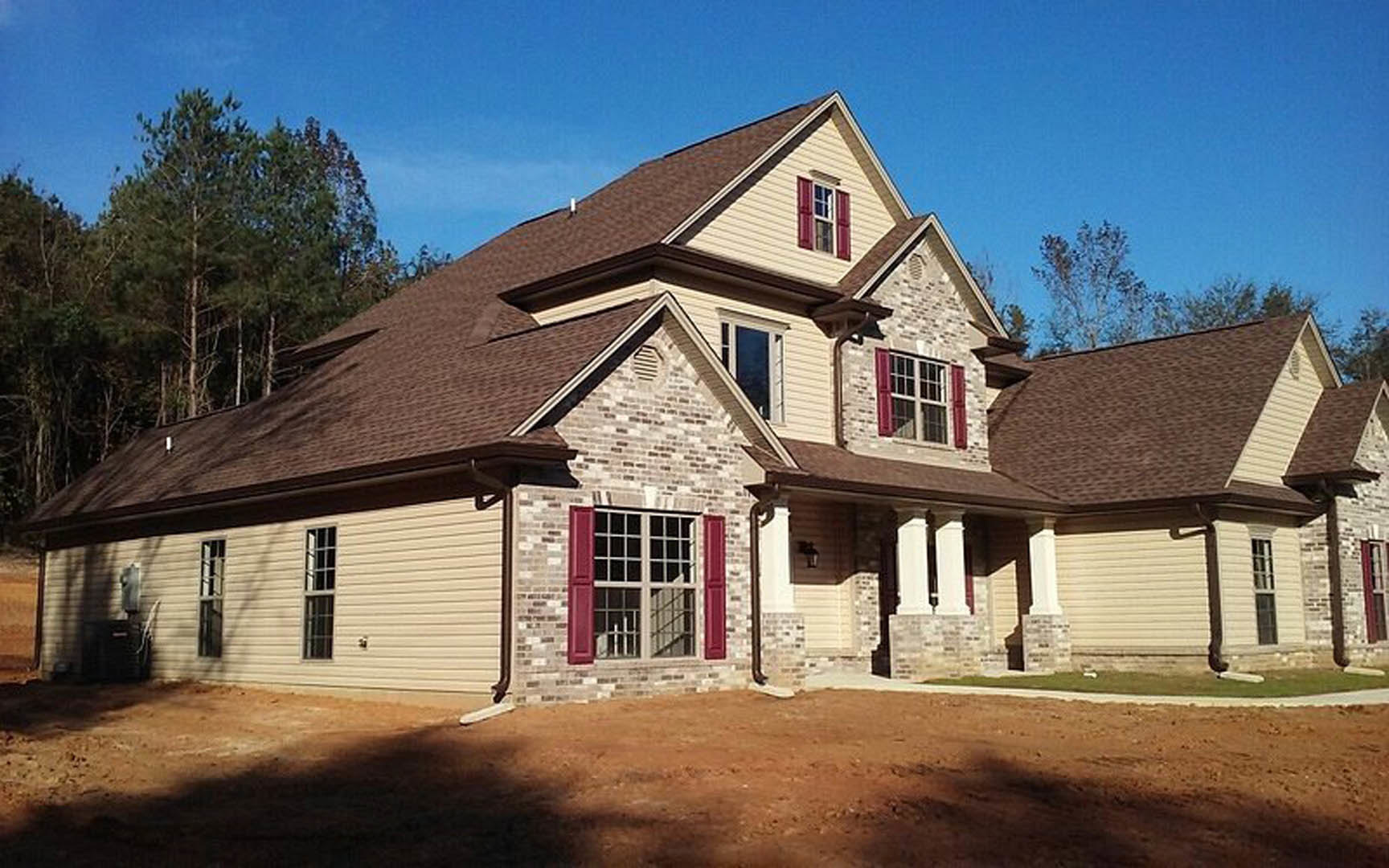 Two-story brick house with brown shingle roof, red shuttered windows, and mature tree in front yard