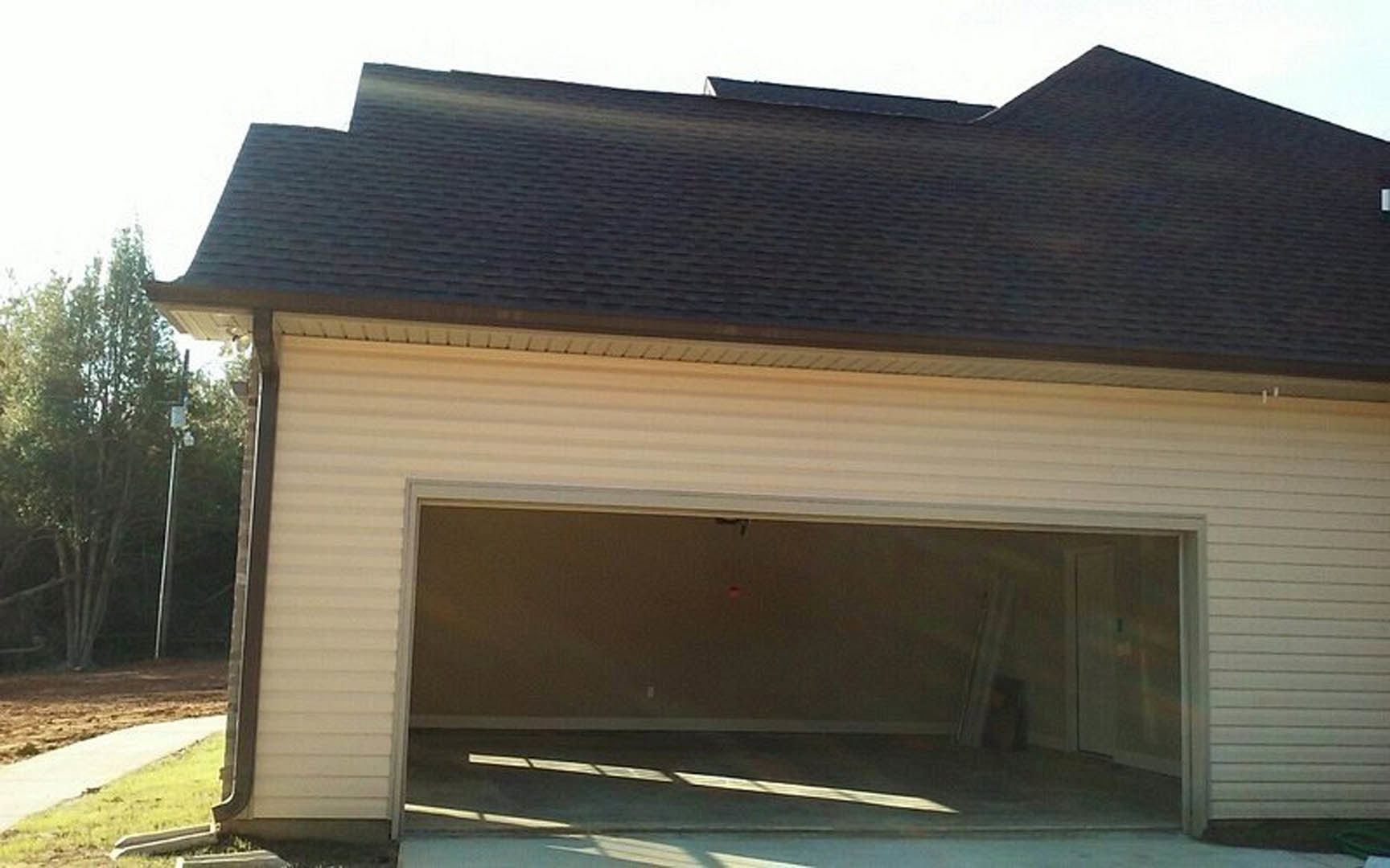 Spacious garage with wide open door, white siding, shingle roof, leafy tree nearby, sunlight streaming onto concrete driveway