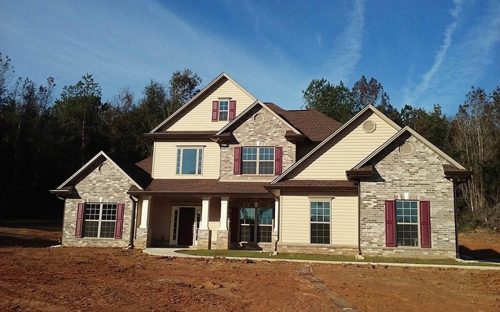 Two-story home with white siding, large multi-pane windows, prominent wooden front door, dirt driveway, and mature trees in the yard under a partly cloudy sky