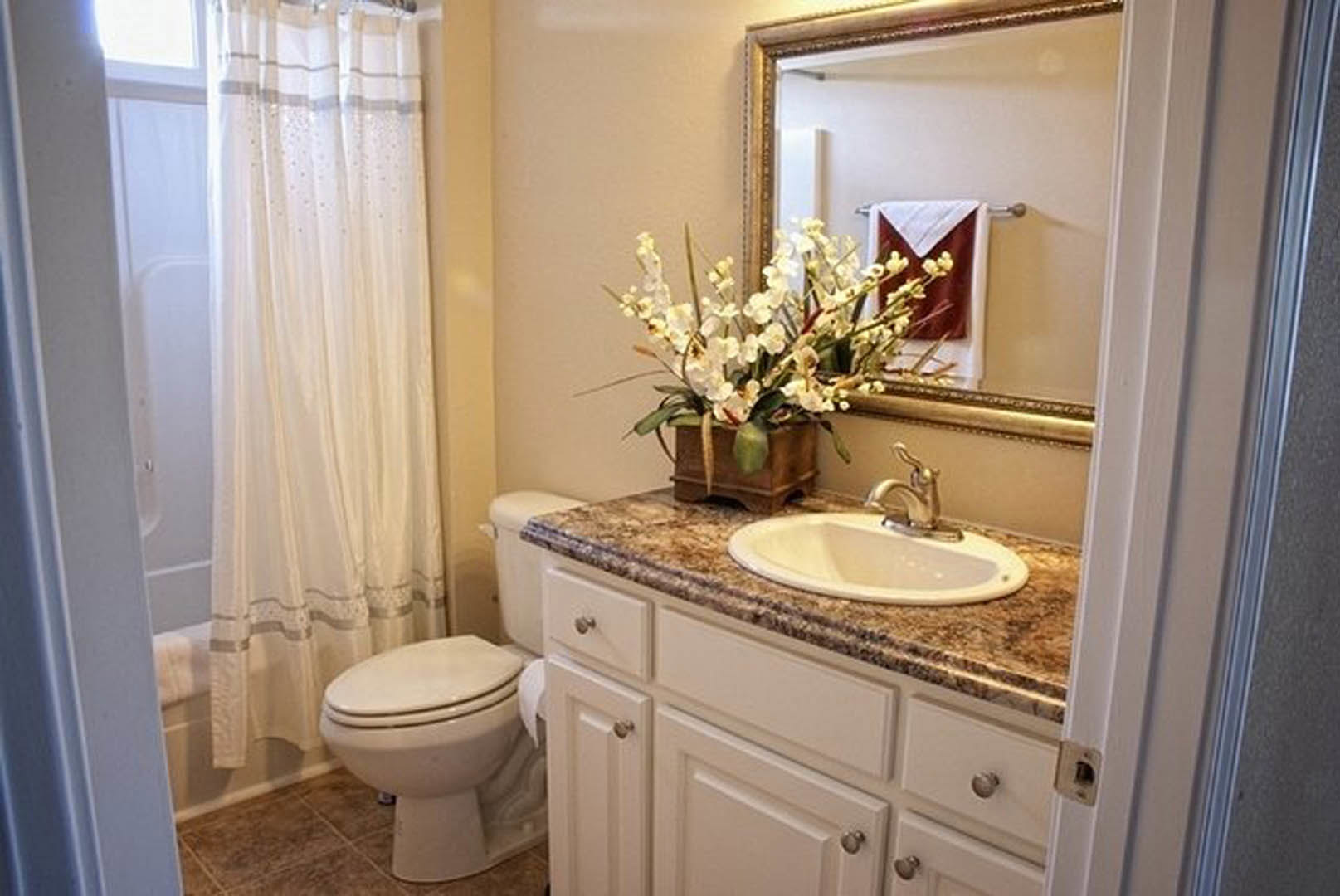 Modern bathroom featuring a rectangular mirror above a white sink with chrome faucet, white cabinetry, tiled walls, a toilet with white lid, and a potted white flower on the