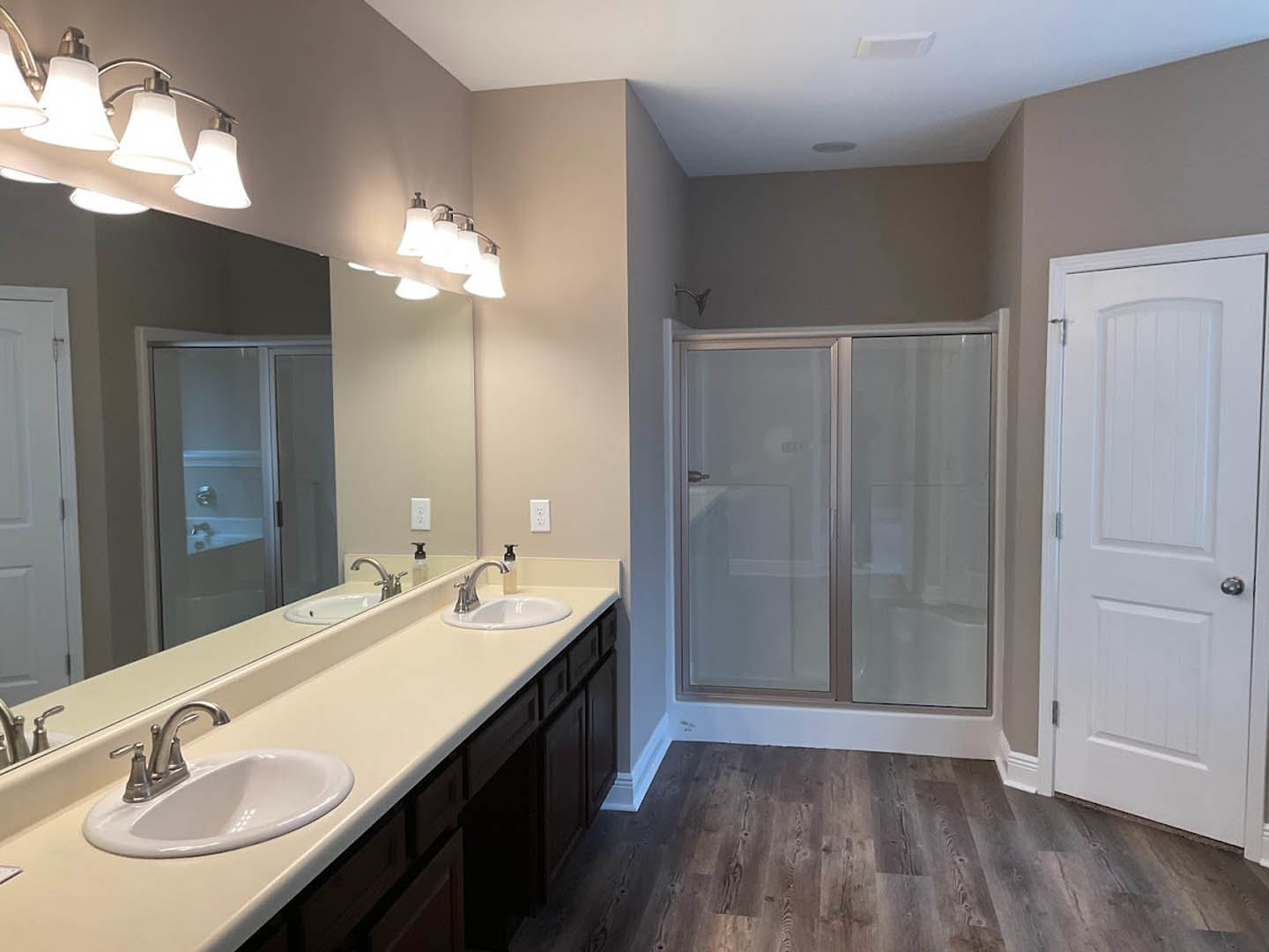 Bathroom with double vanity featuring undermount sinks, quartz countertop, glass-enclosed shower with tile walls, large mirror above sinks, white cabinetry, and row of modern light