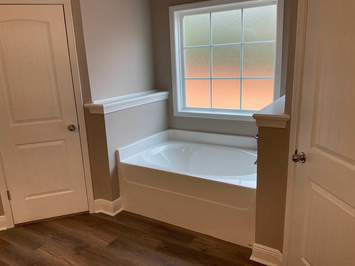 Freestanding white bathtub on wood floor beside frosted glass window, white door with silver handle, light reflecting off tub surface