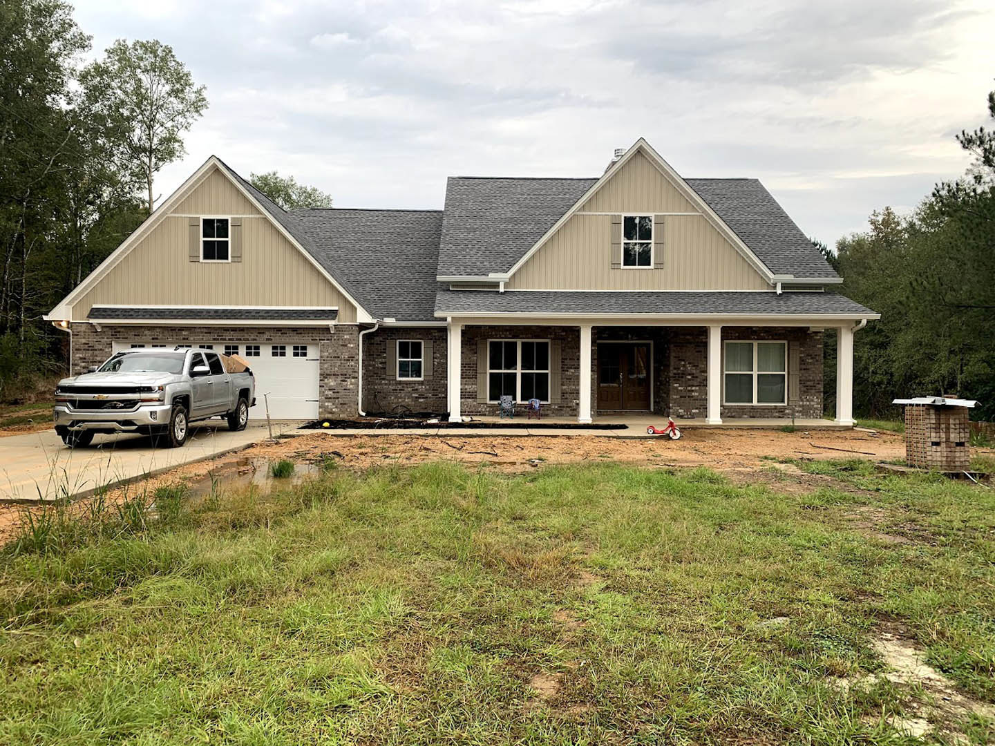 Brick and stone exterior home with double glass panel doors, silver truck parked in driveway, grassy yard, train visible in background, cloudy sky overhead