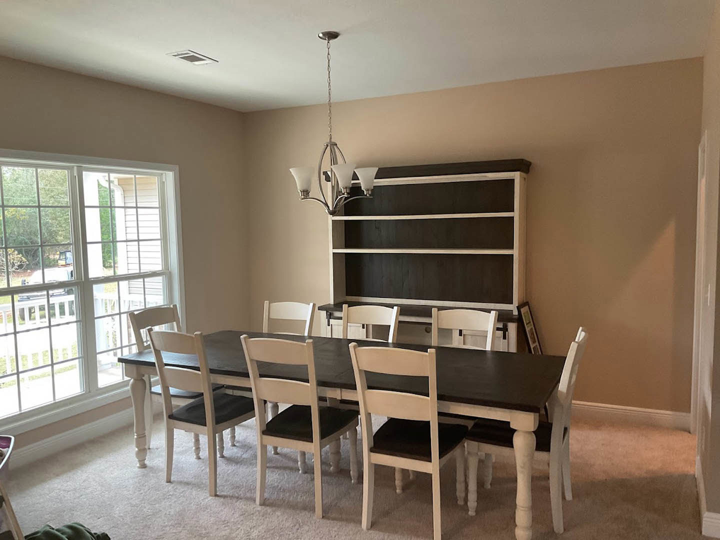 Wood dining table with upholstered chairs on hardwood floor, large window with white trim, built-in shelving along wall, neutral paint, recessed ceiling lights