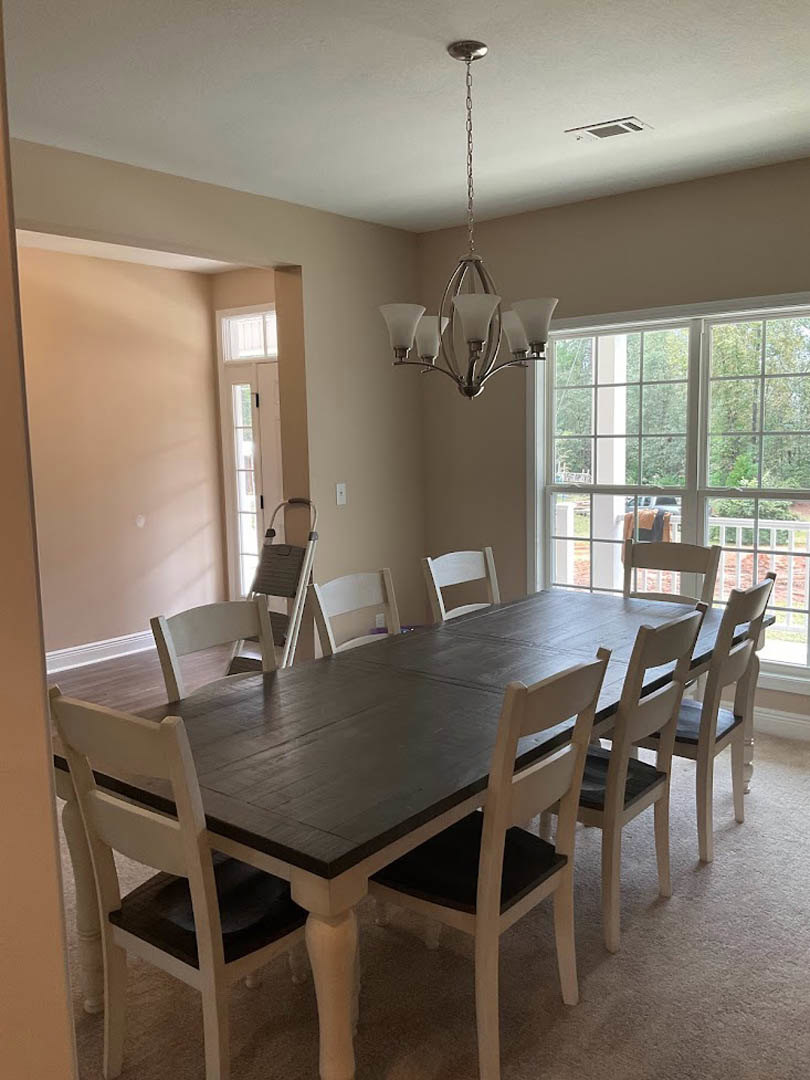 Wood dining table surrounded by upholstered chairs, five-light chandelier hanging from chain above, large window with white trim, neutral walls, hardwood floor