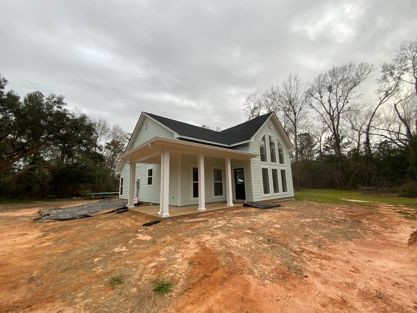 Wood-framed house under construction with covered porch, exposed plywood walls, dirt patch and large grey rock in foreground, leafy trees in background