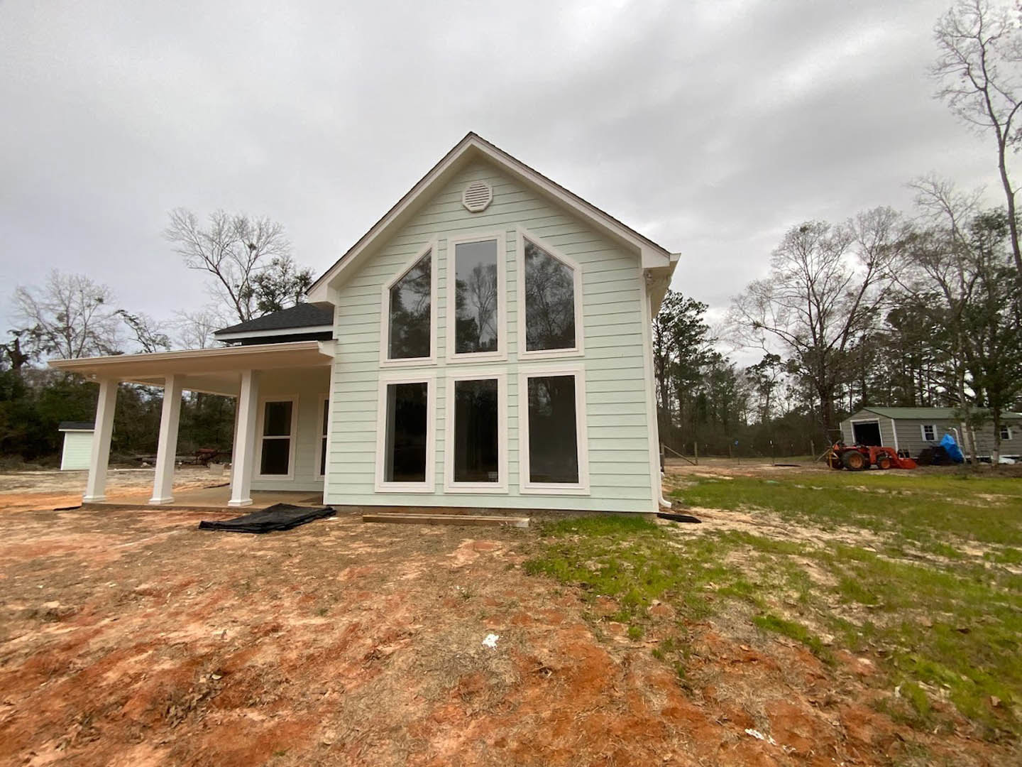 Modern home under construction with exposed framing, multiple large windows, light-colored siding, tractor parked on snowy lawn, leafless tree in background, overcast sky