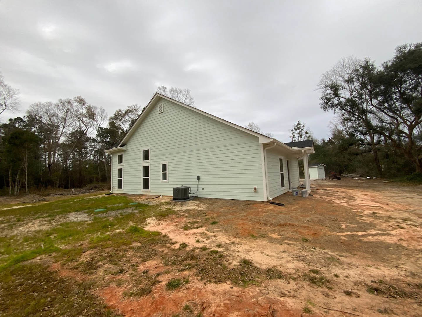 Modern cottage-style home with light-colored siding, dark roof, and large central black feature; surrounded by dirt field, mature trees, and open rural landscape.