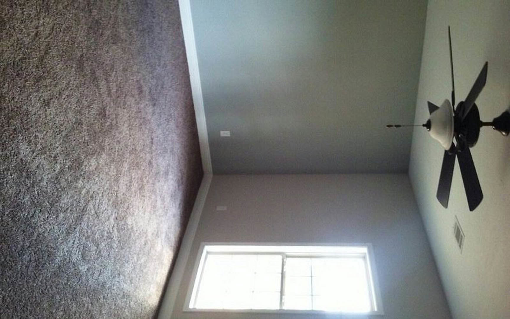 Neutral-toned carpeted room with white walls, large window letting in natural light, and ceiling fan with integrated light fixture