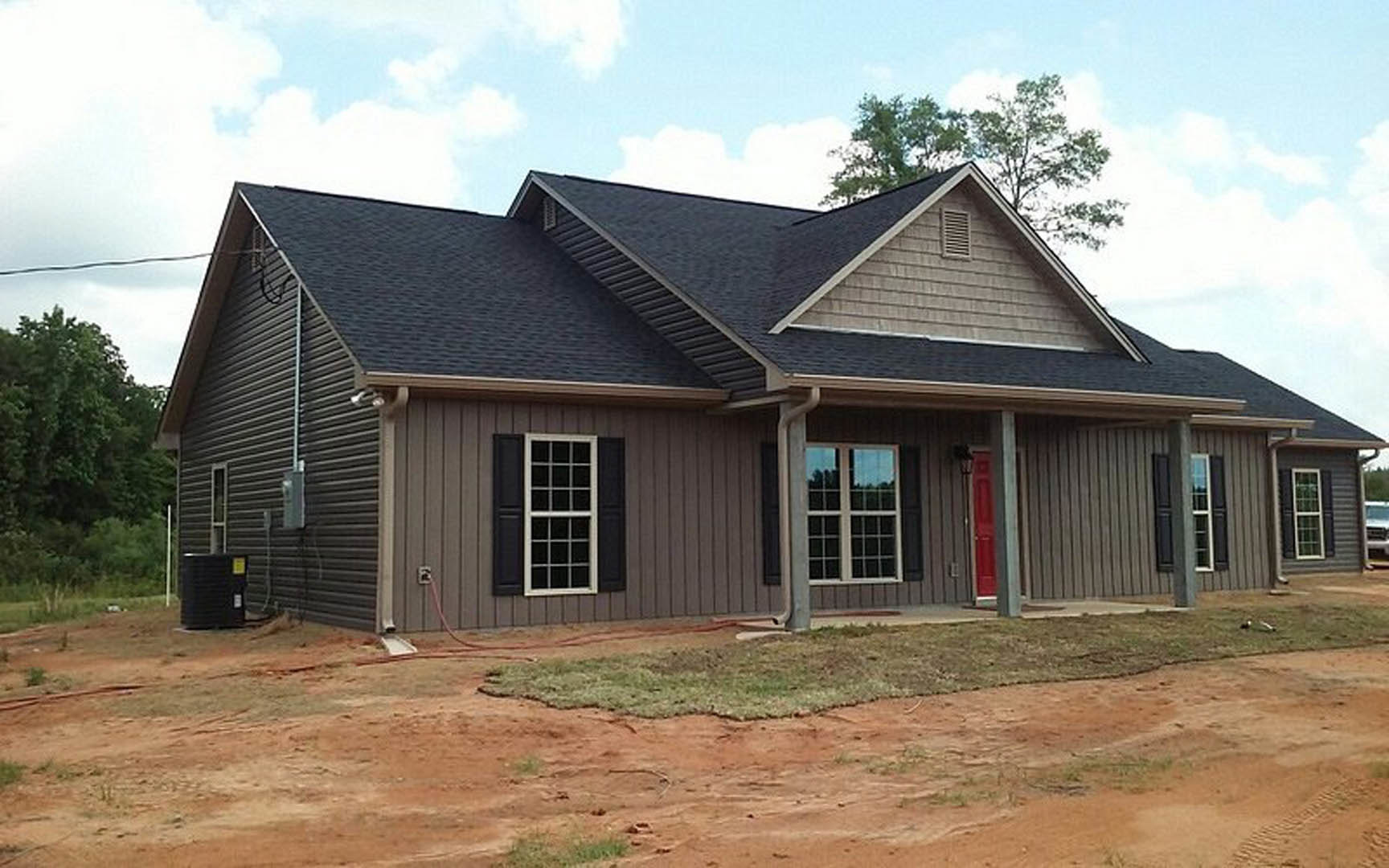 Red front door on a cottage-style home, white-trimmed window, gray shingle roof, dirt patch near foundation, trees and cloudy sky in background