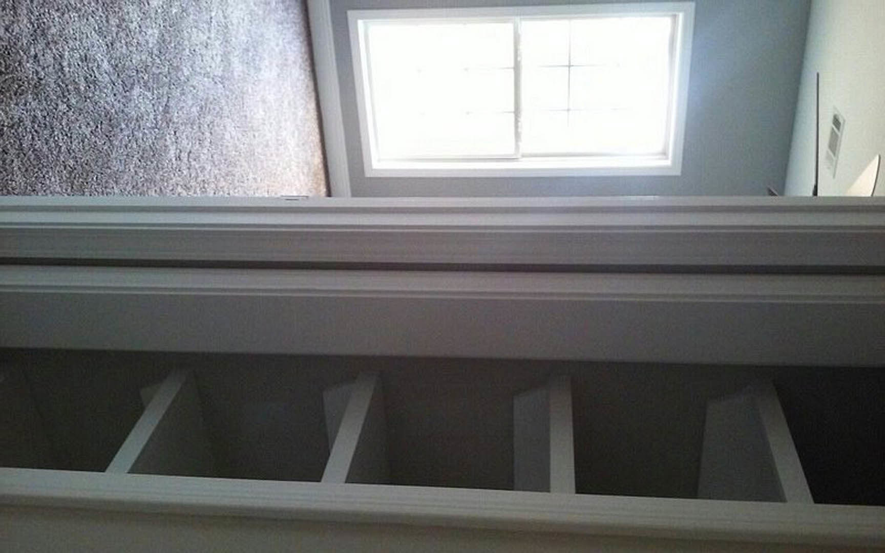 White-framed window with closed blinds set in a white wall, adjacent to a white door, grey carpet flooring, and a wall vent; black stripe detail visible on the wall.