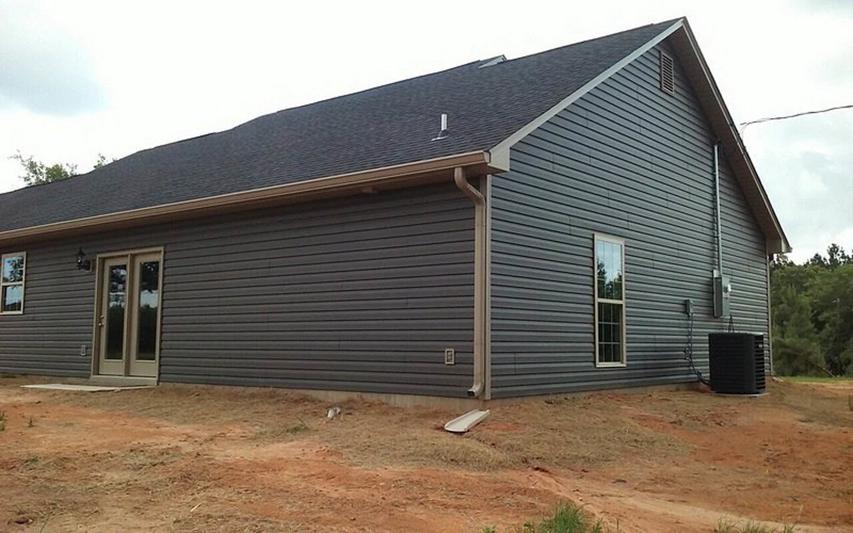 Grey siding house under construction with visible gutter, close-up door, black rectangular object on table, windows reflecting trees, outdoor setting with roof and surrounding