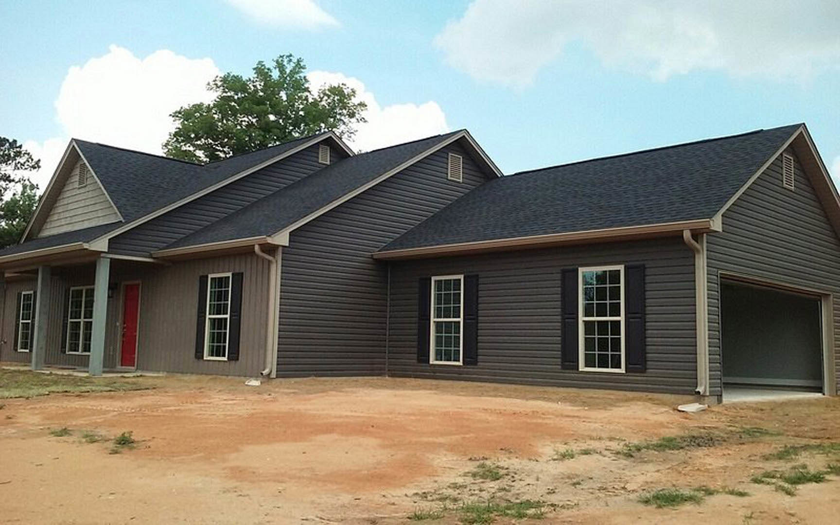 Two-story home with white-trimmed multi-pane windows, gray siding, and a dirt driveway, surrounded by green-leaved trees under a cloudy sky