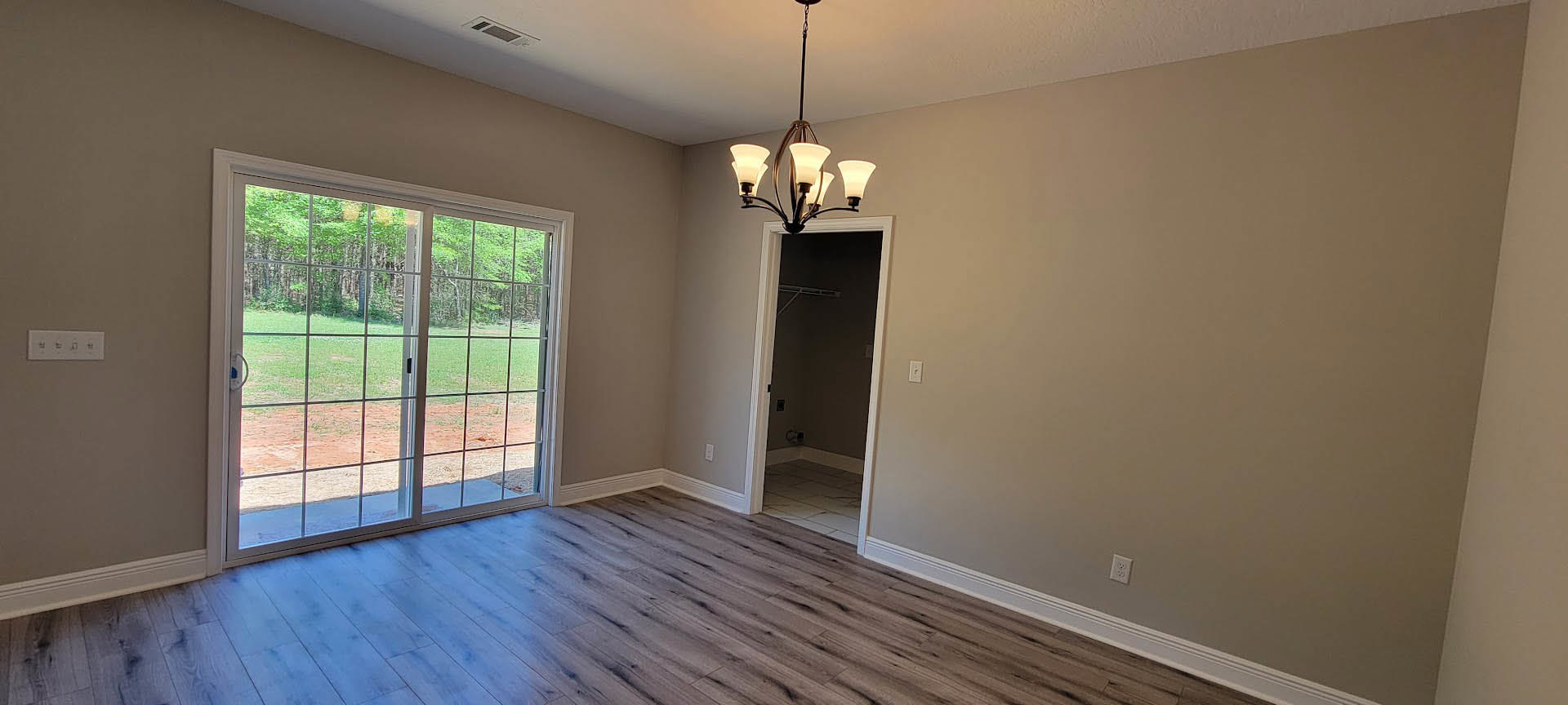Wood flooring with white trim, sliding glass door overlooking grassy field, traditional door with silver handle, modern chandelier, white thermostat with black buttons on plaster