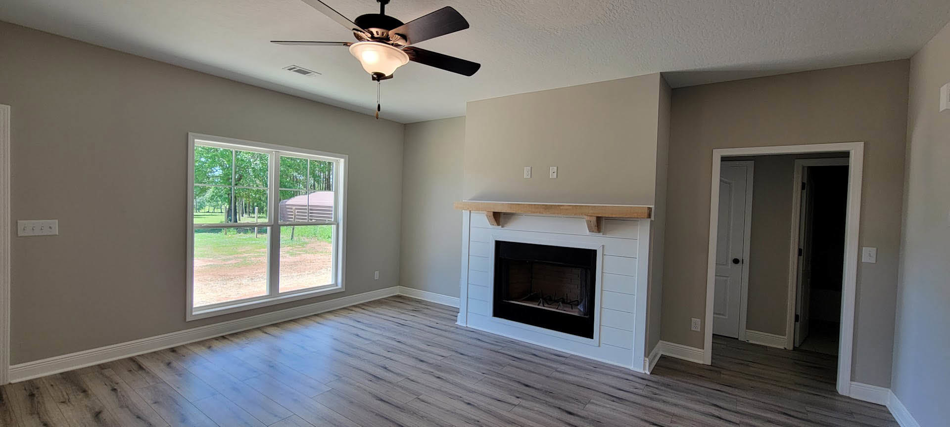 Living room with black-framed fireplace, ceiling fan with light, large window overlooking field and trees, wood flooring, and white wall panel with knobs