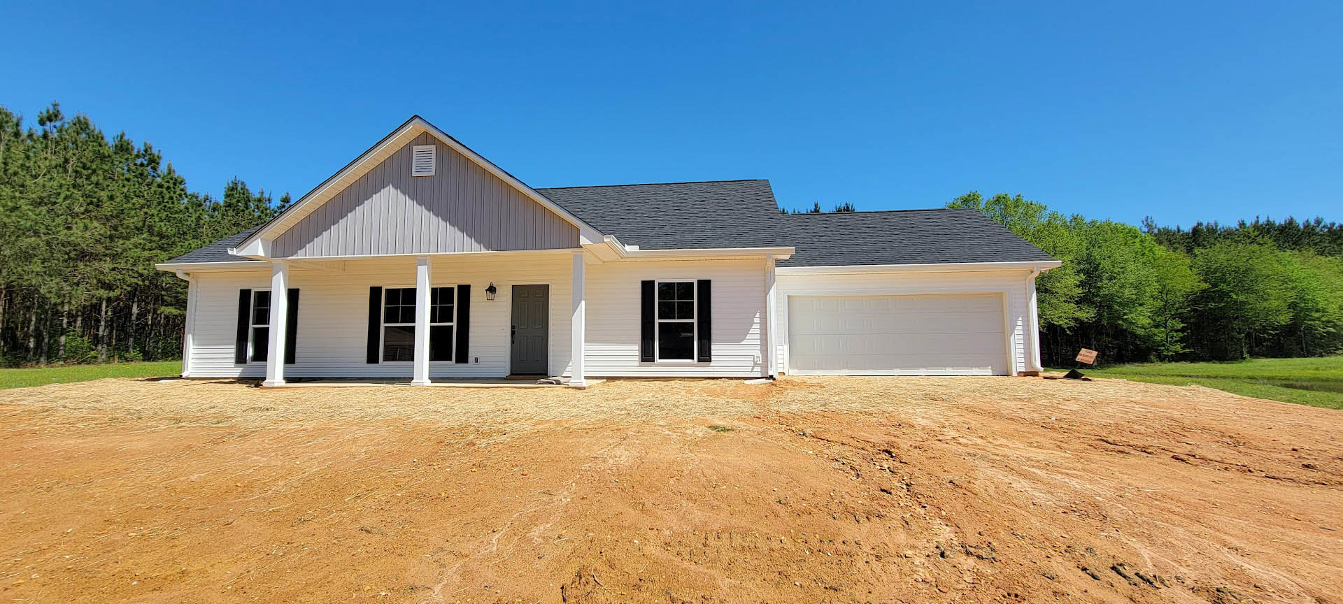 White garage door with black trim on a modern house, black shingle roof, dirt driveway, blue sky overhead, trees in background.