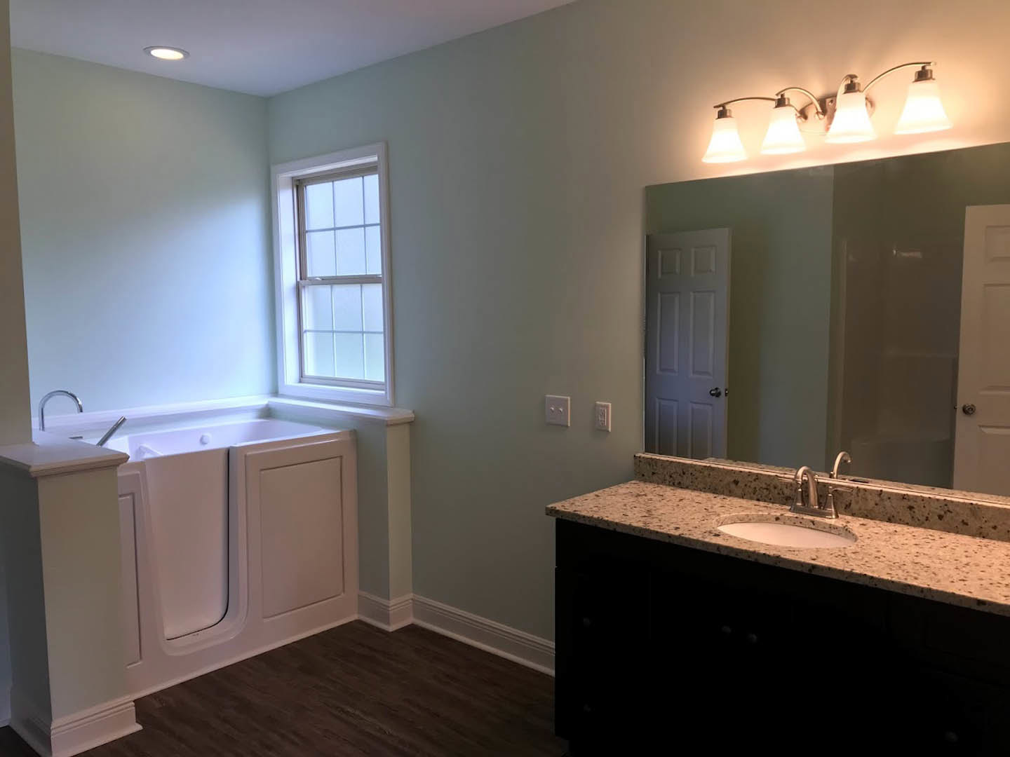 Bathroom with freestanding tub, white sink set in stone countertop, brown-framed window, white door with black handle, light fixture with two lamps, neutral tile flooring, and