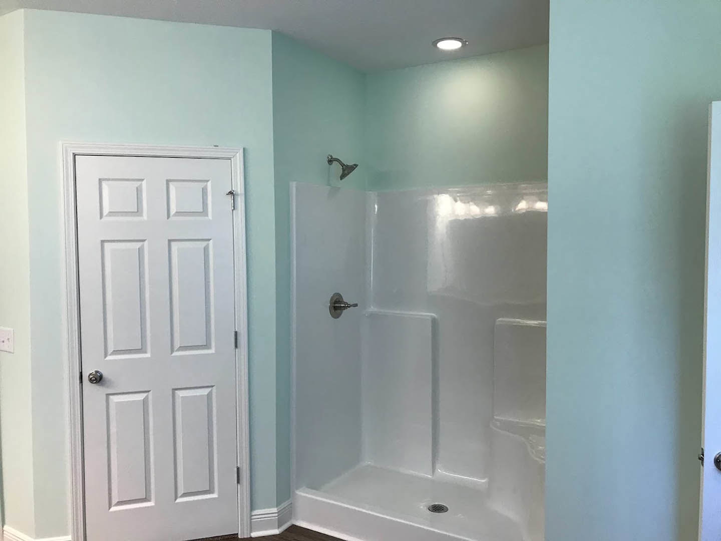 White bathroom with glass shower enclosure, silver shower head and handle, white door with silver knob, light-colored walls and ceiling, close-up of chrome faucet.