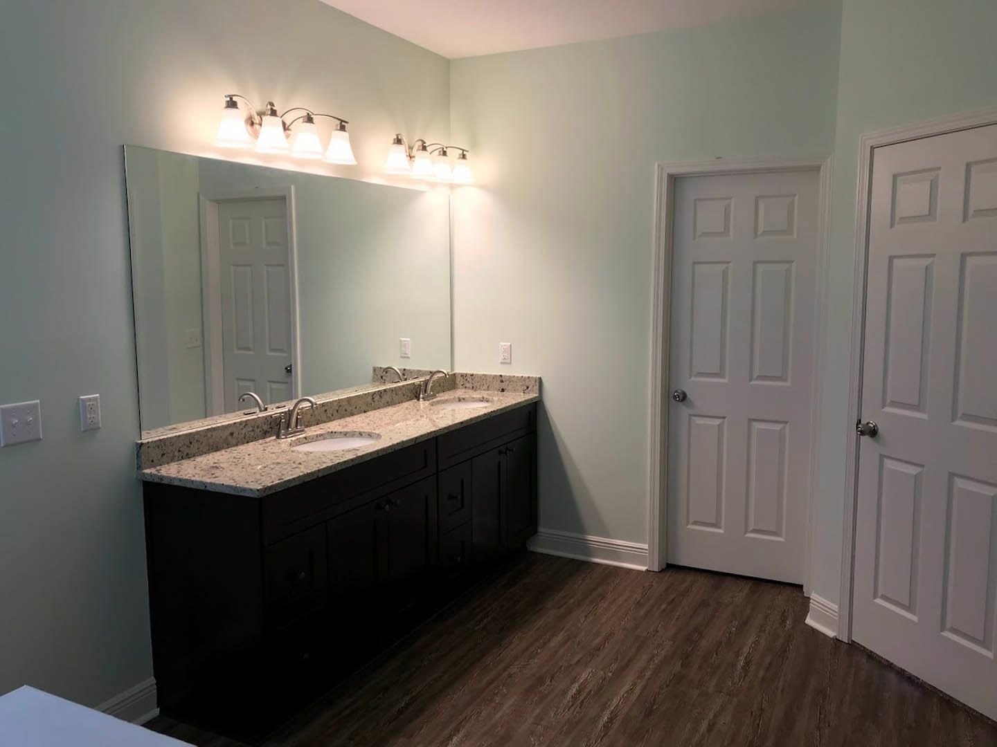 Bathroom with double vanity featuring white sinks, large wall mirror, light fixture with three lamps above, white cabinetry, tiled floor, white door with silver knob, and white