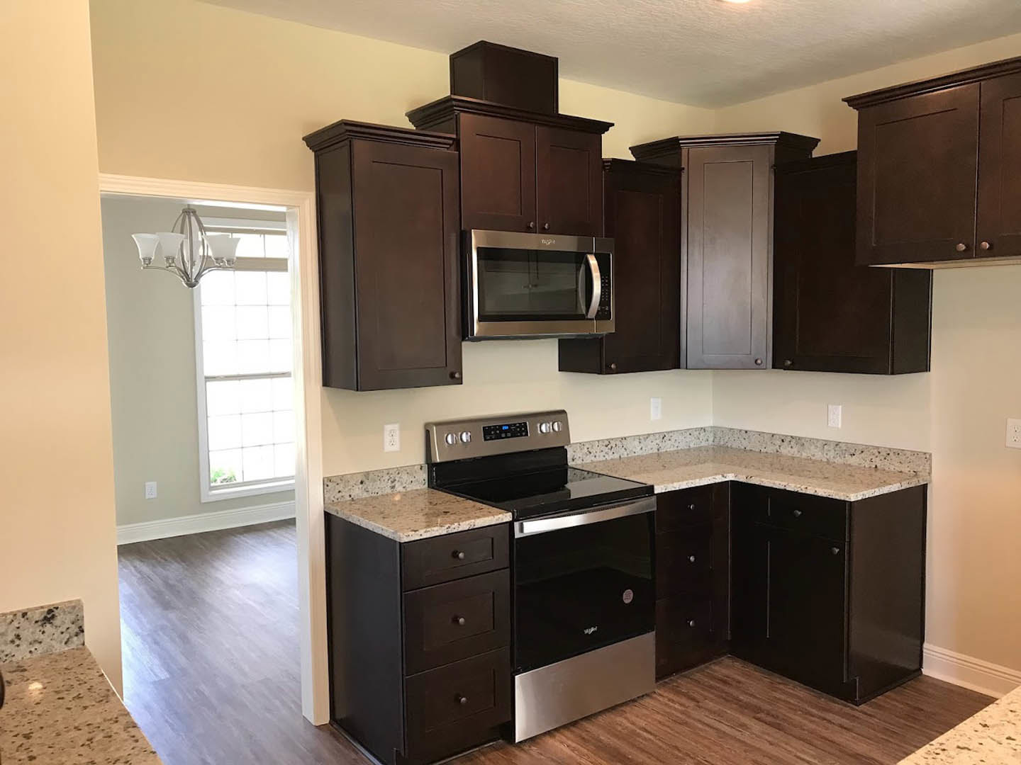 Dark wood kitchen cabinets, granite countertops, stainless steel microwave and stove, modern light fixture, white door, black and silver computer on counter