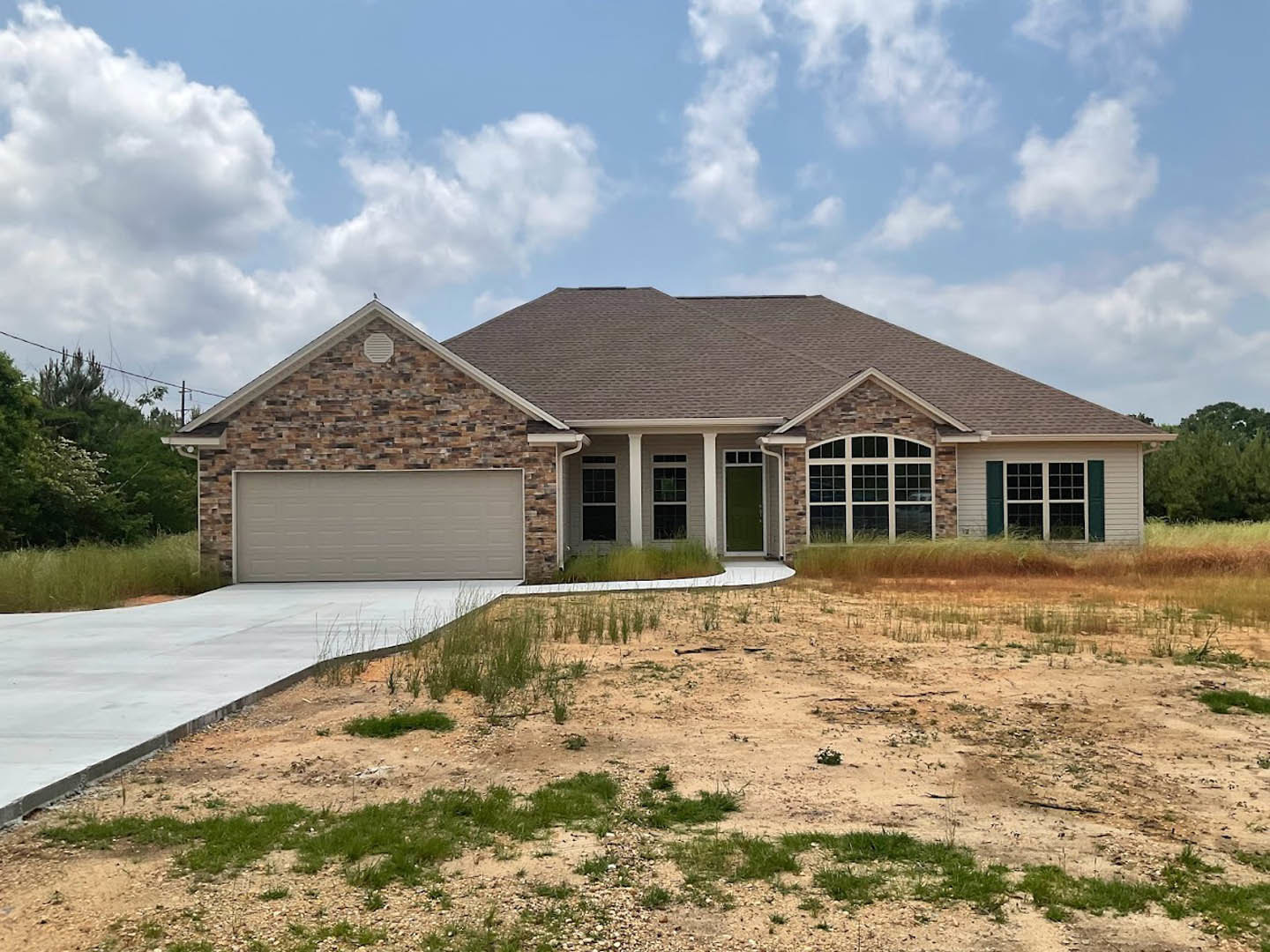 Two-story house with stone and brick exterior walls, white garage door, green front door with white trim, paved driveway, patchy grass and dirt yard, gray shingle roof with