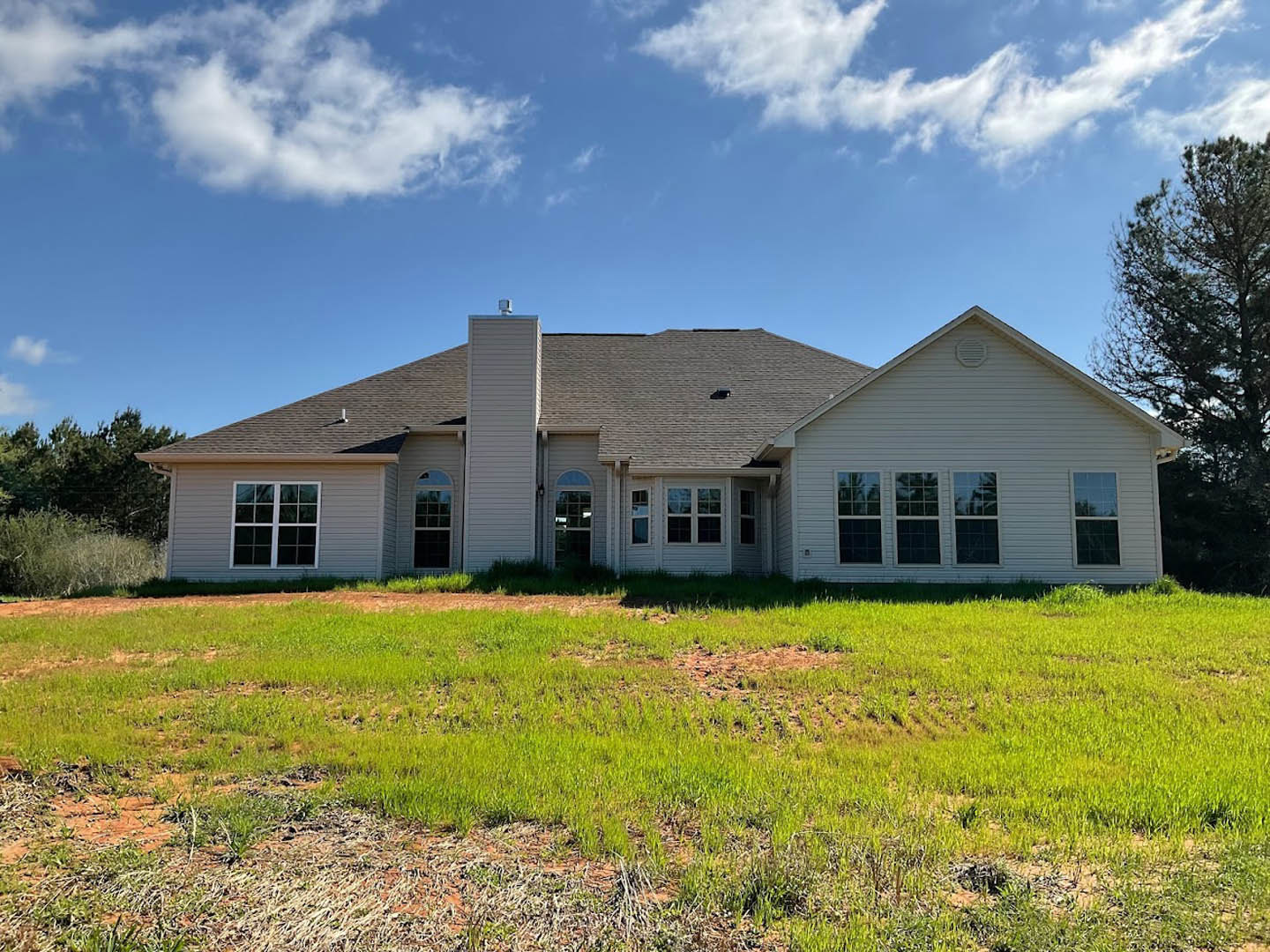 White farmhouse with multi-pane windows, large front lawn, mature trees, and blue sky with scattered clouds