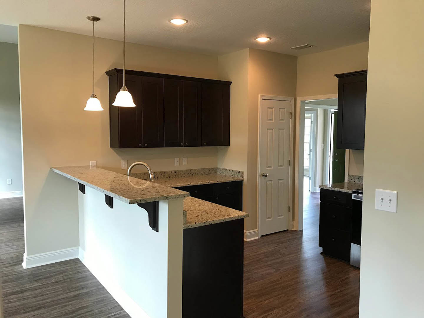 Open kitchen with wood flooring, white cabinetry, bar seating, stone countertop with stainless steel sink, white door with silver handle, wall outlet and switches, white walls