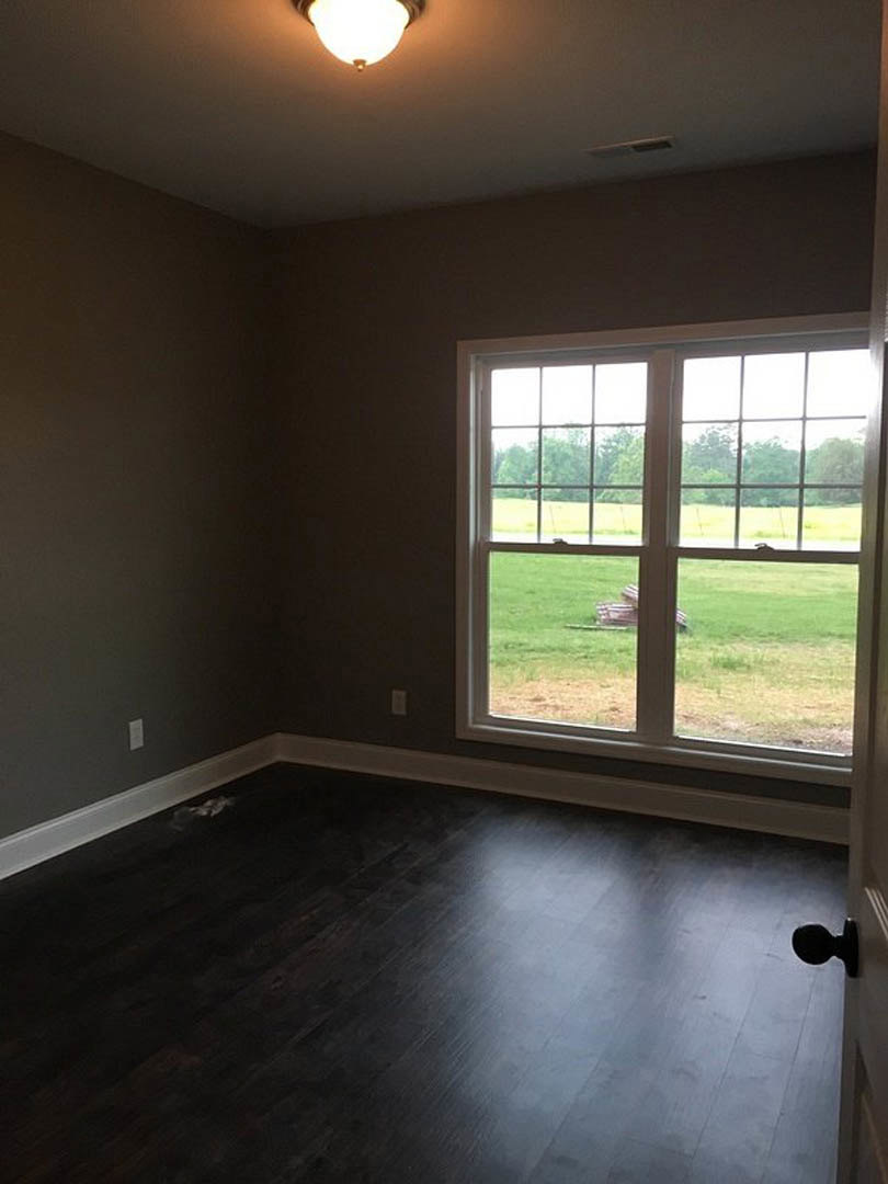 Dark wood flooring in a bright room with a large window overlooking a grassy field, white walls, and a black door knob on a white door