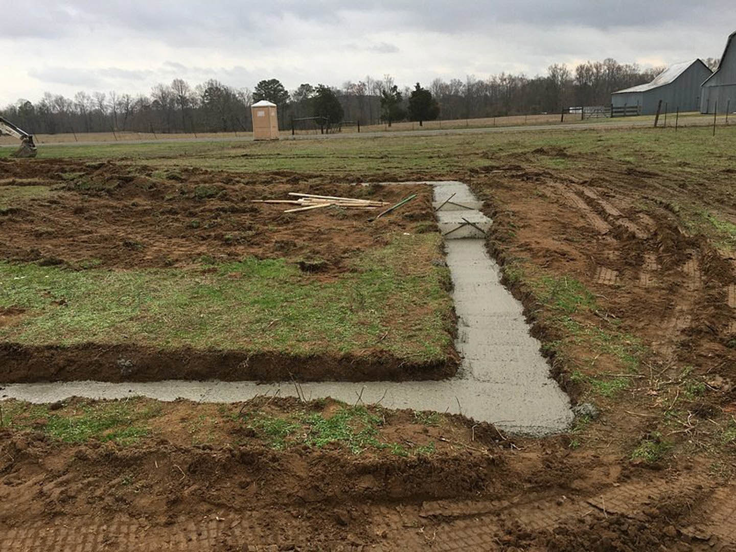 Concrete drainage ditch running through grassy field with patches of snow, surrounded by soil and sparse trees under cloudy sky
