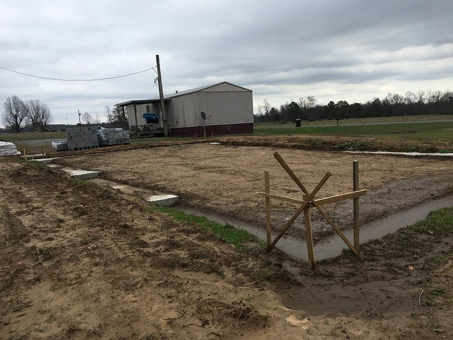 Dirt field bordered by a wooden fence, leafless trees, stone, and a distant building with satellite dishes under a cloudy sky
