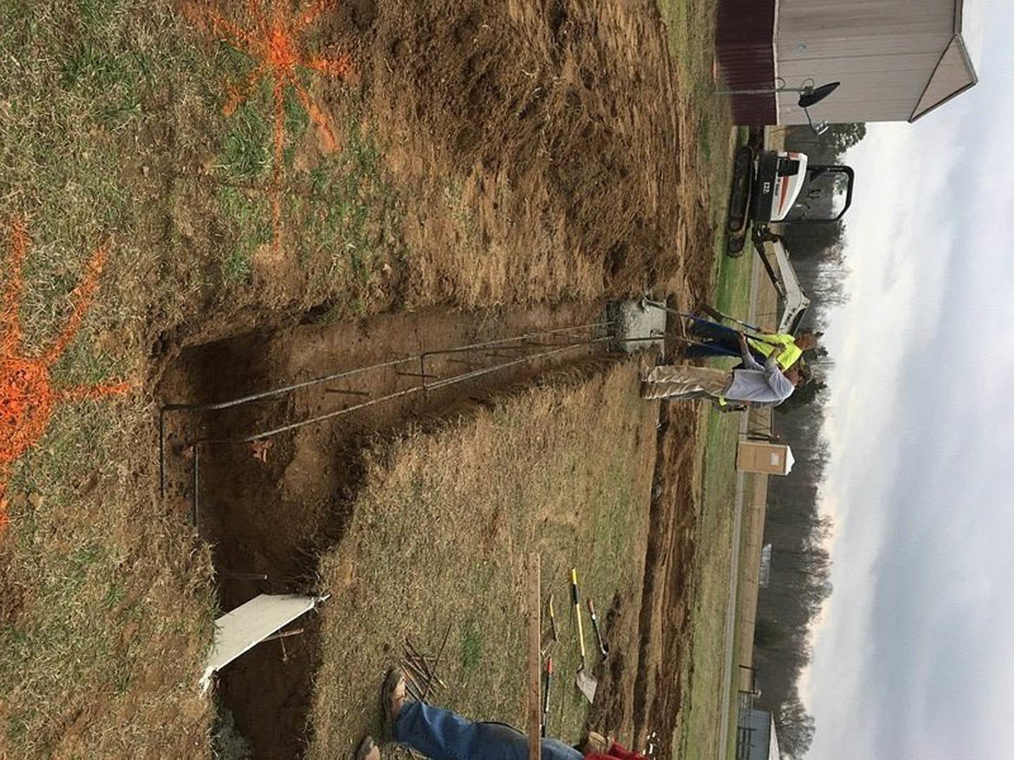 Two men standing in a grassy field, one wearing a yellow safety vest and holding a stick, with plants and a satellite dish visible nearby.