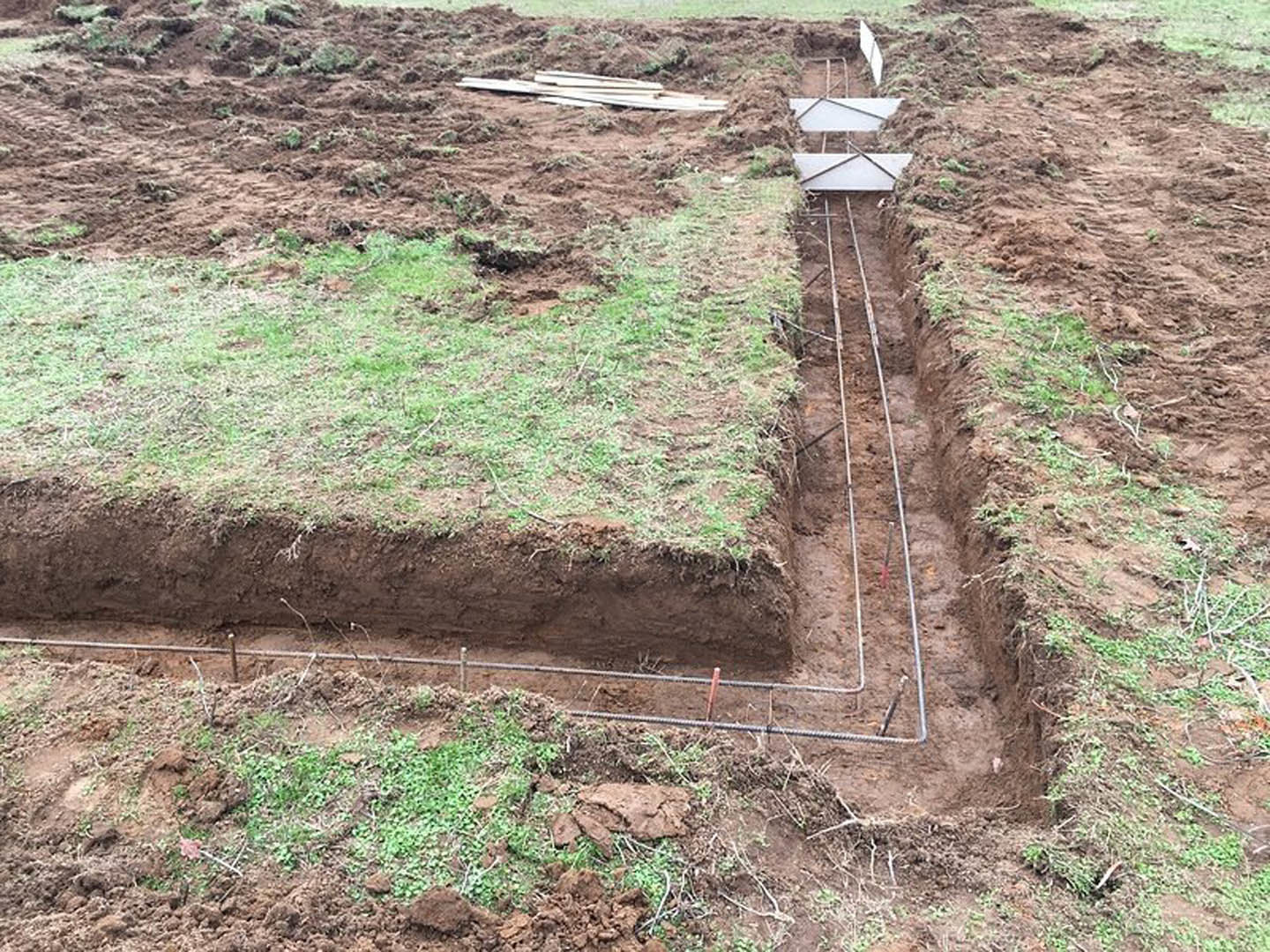 Open trench with exposed pipes running through soil, surrounded by patches of grass and metal bars on a dirt field