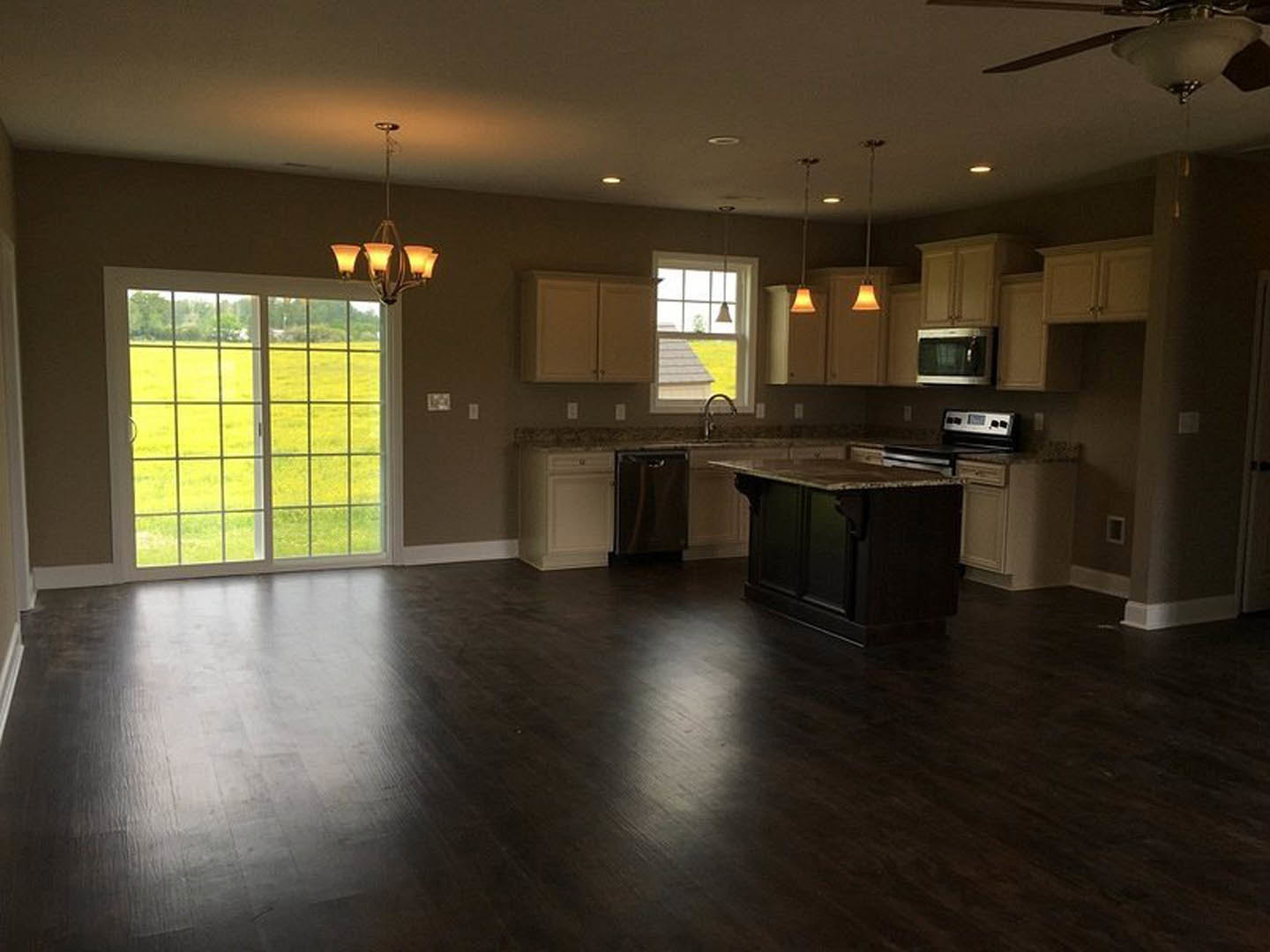 Kitchen with large window, black marble-topped island, dark wood flooring, white walls, stainless steel refrigerator, microwave oven with handle, sliding glass door overlooking