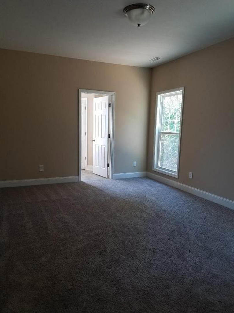 Carpeted room with a white door open, silver handle, white-framed window, ceiling light fixture, and neutral plaster walls