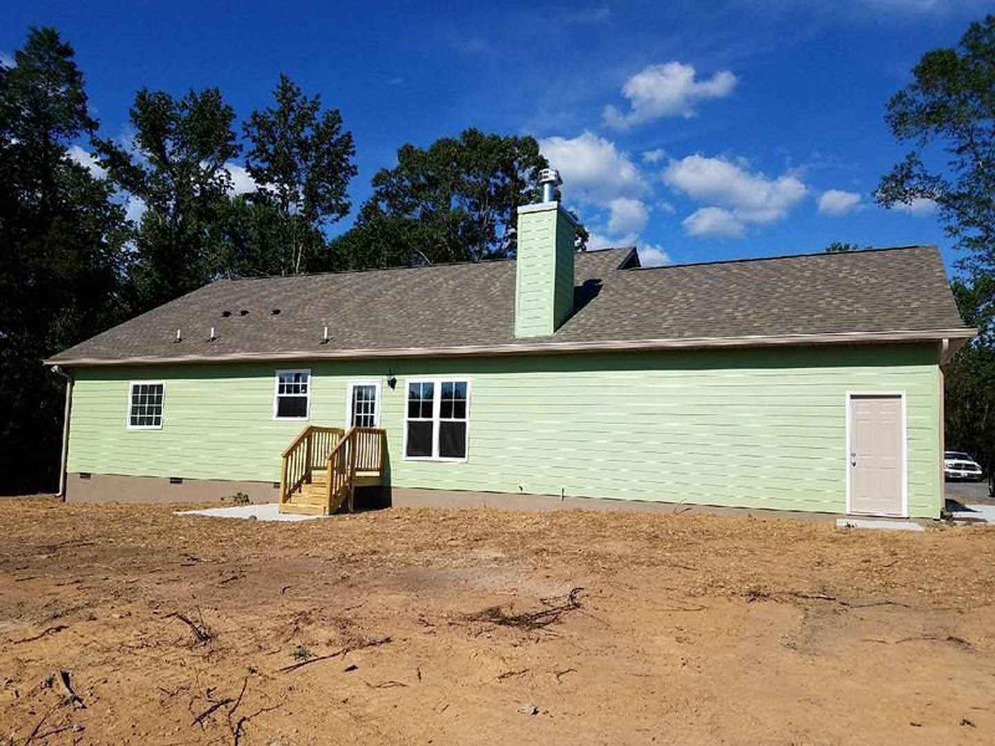 Green house with white trim, wooden porch and stairs, white door with silver handle, dirt yard scattered with sticks, blue sky and clouds overhead