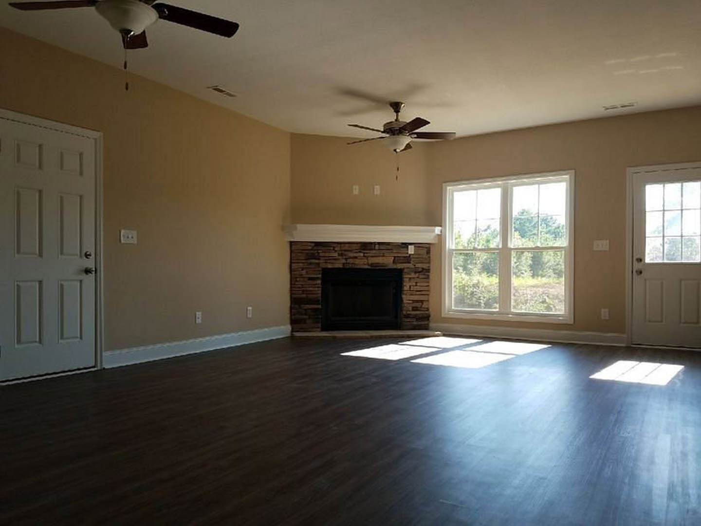 Living room featuring wood flooring, stone fireplace with white mantel, ceiling fan with light fixture, large window overlooking trees, and white door with glass insert.
