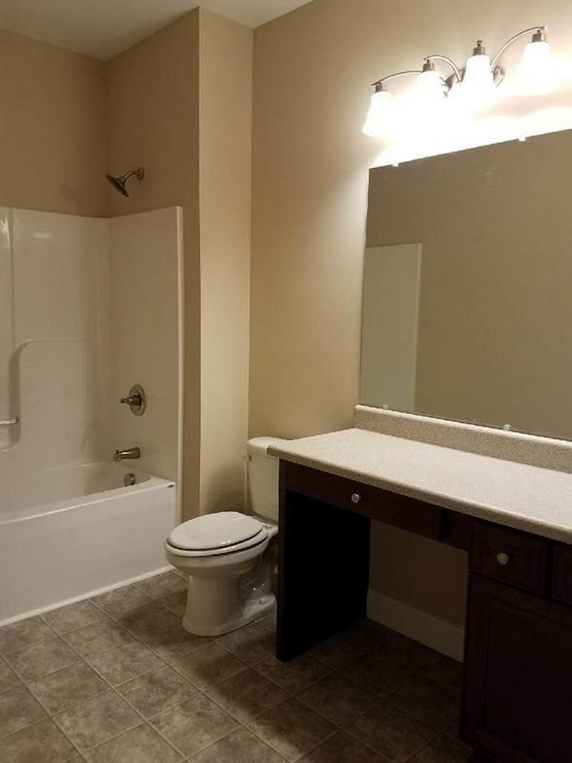 Bathroom with white tile floor, white bathtub beside closed toilet, single sink vanity, three-light fixture above mirror, wall-mounted shower head
