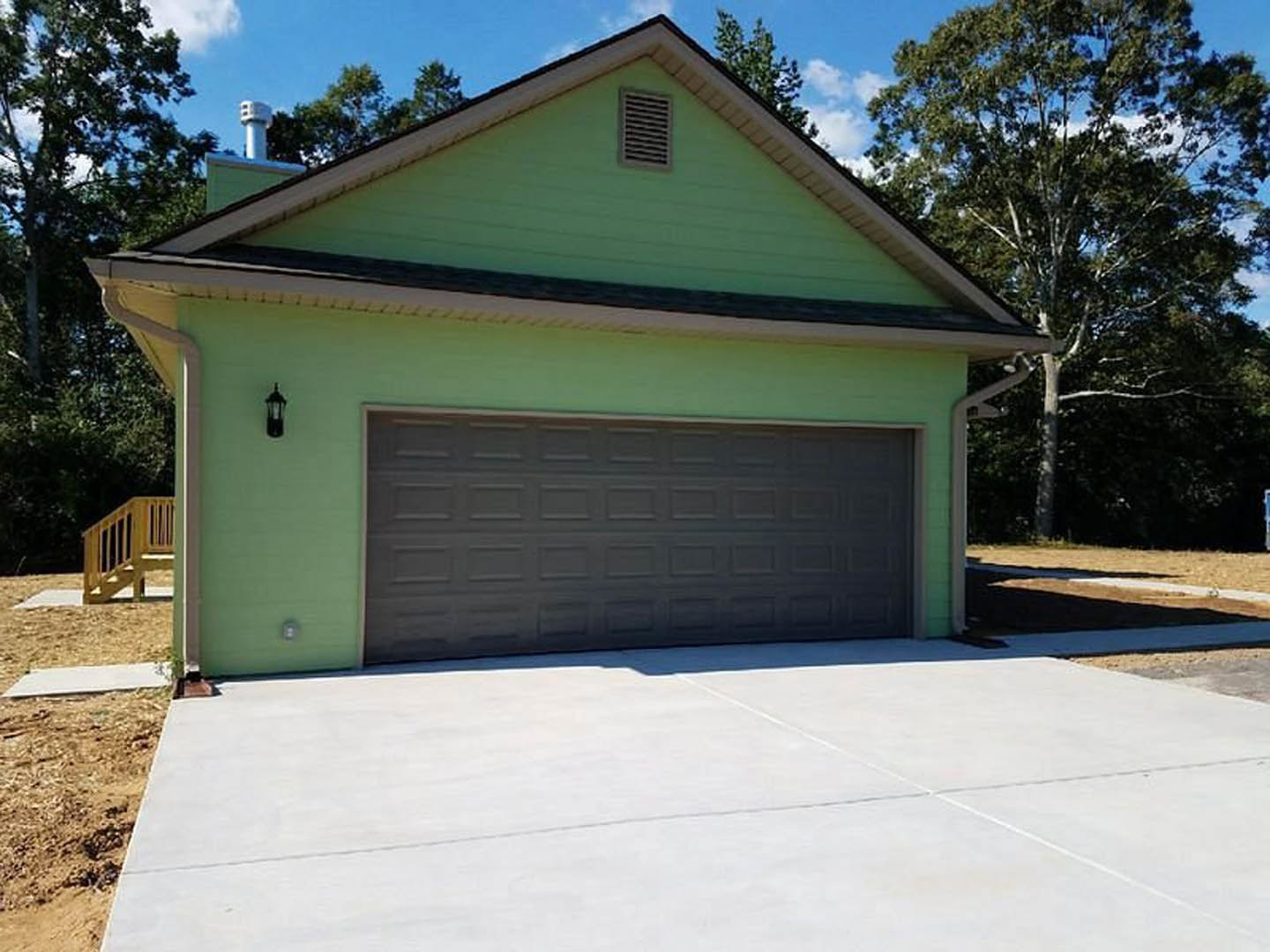 Green house with horizontal siding, attached garage featuring a white paneled door, wooden deck with railing, concrete driveway, leafy trees in background, wall vent visible near