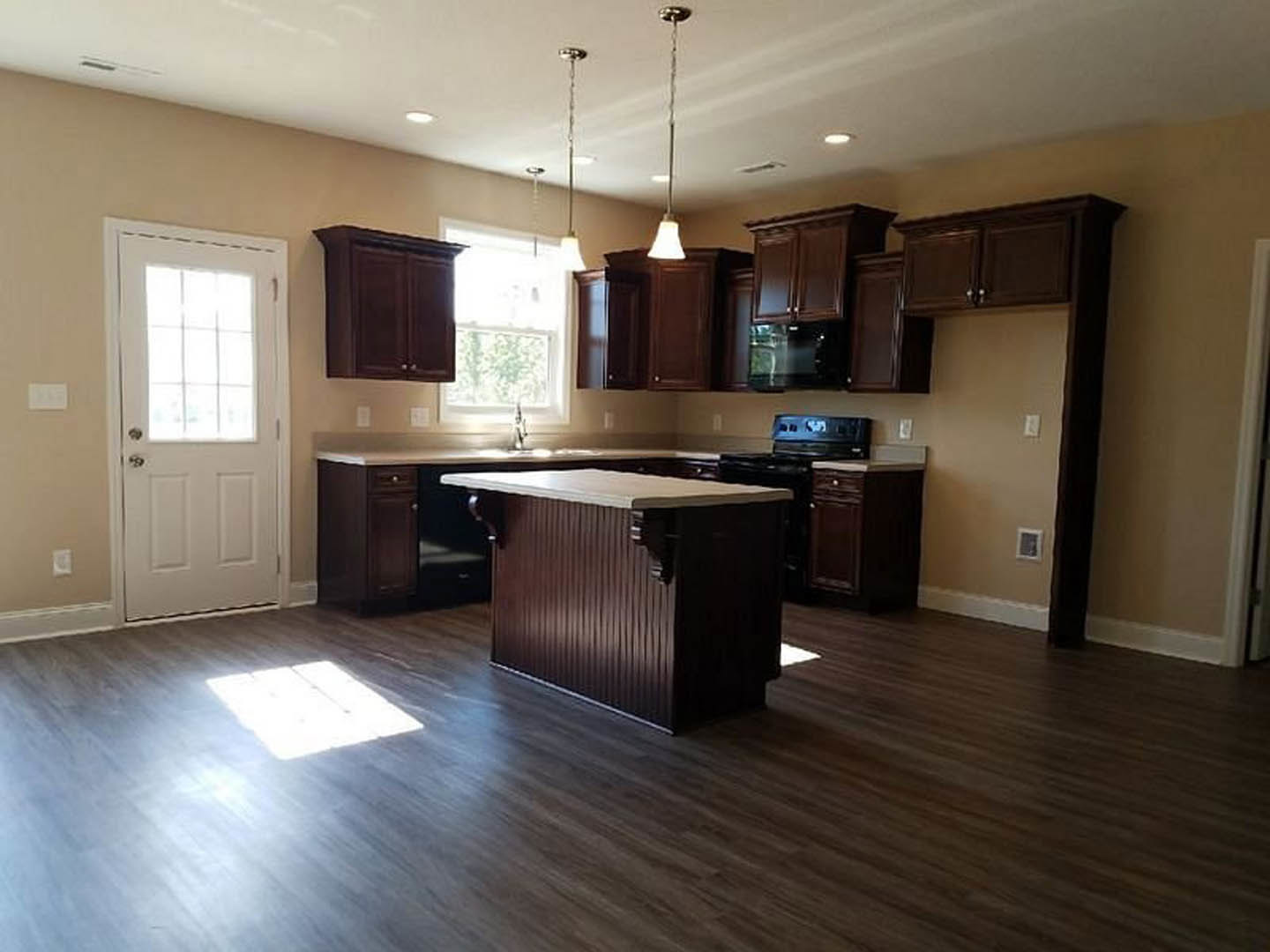 Kitchen with dark wood cabinets, brown and white countertop, black oven, white door with glass window, wooden flooring, overhead lighting, blurred person in background.