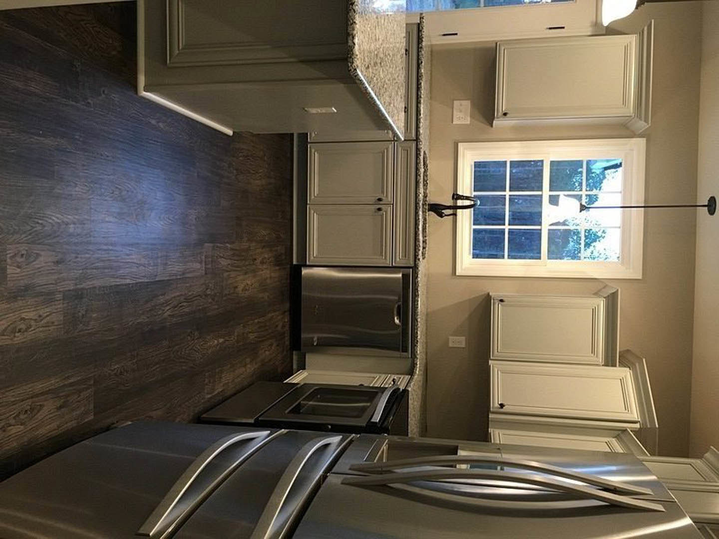 Stainless steel refrigerator and oven beside black cabinetry, dark wood flooring, white-framed window, white ceiling light, and tongs resting on a stainless steel countertop
