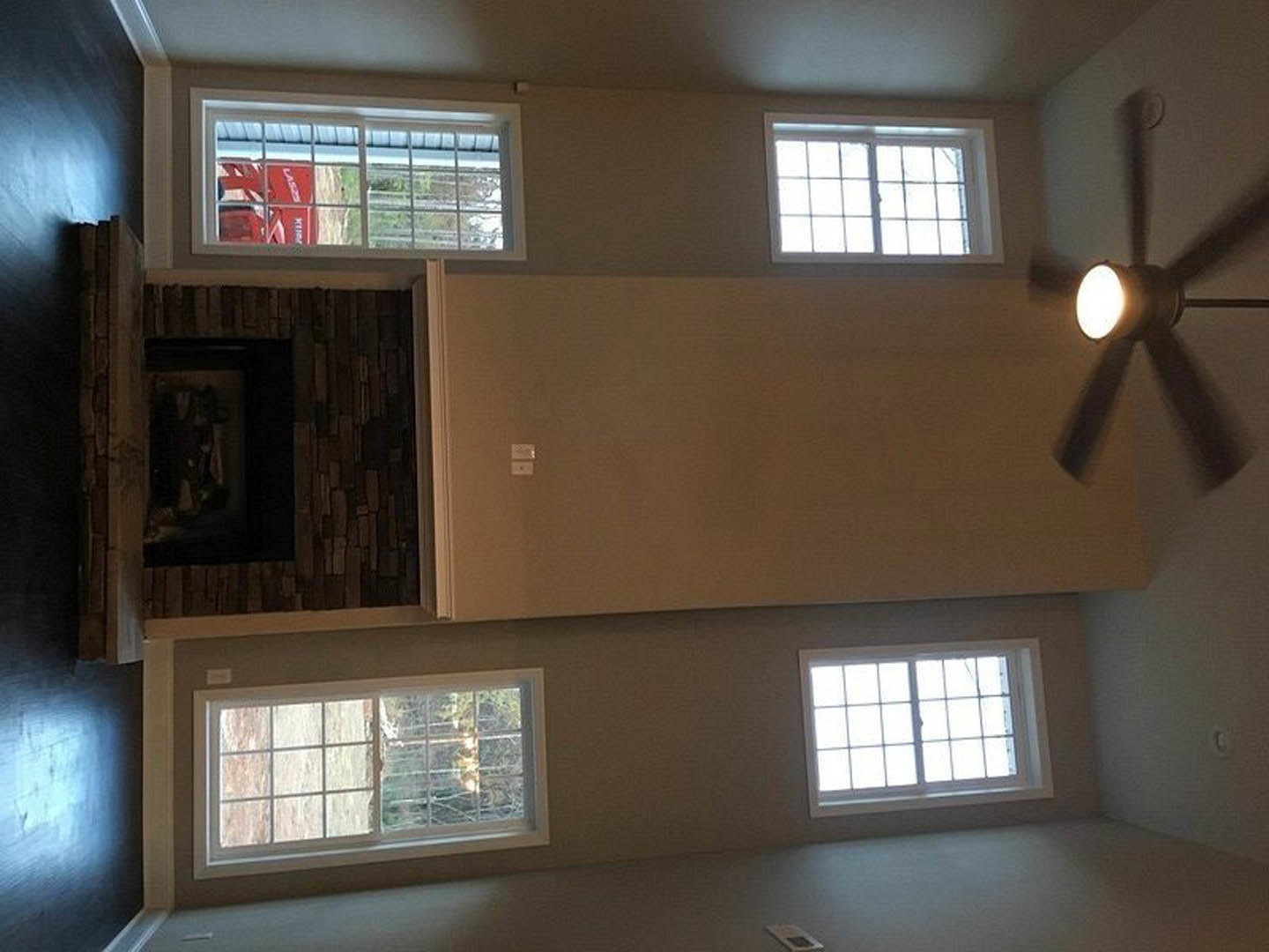 Living room with white plaster ceiling, black and white ceiling fan with light, stone fireplace, large grid-style windows, and daylight streaming in.